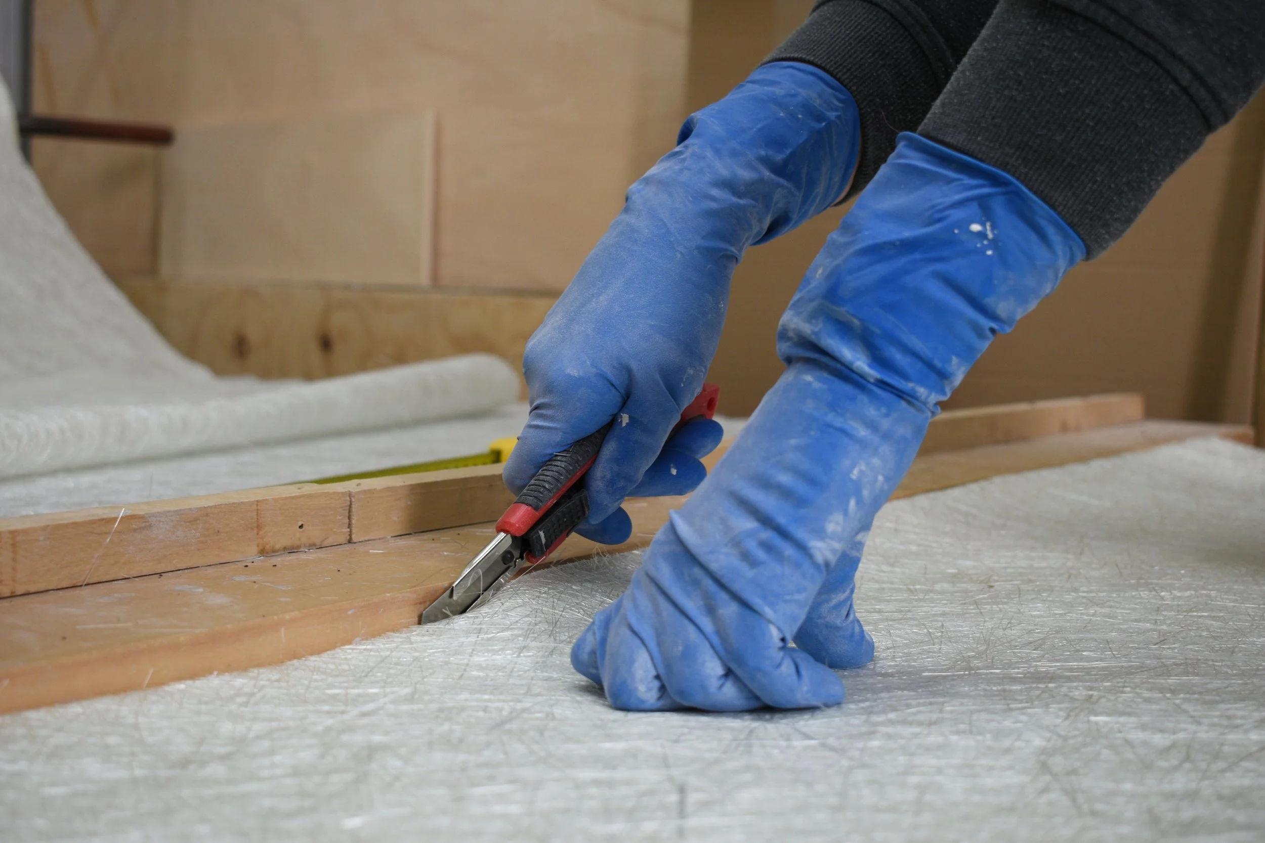 Person wearing blue gloves using a utility knife to cut a piece of fiberglass mat on a wooden table in a workshop.