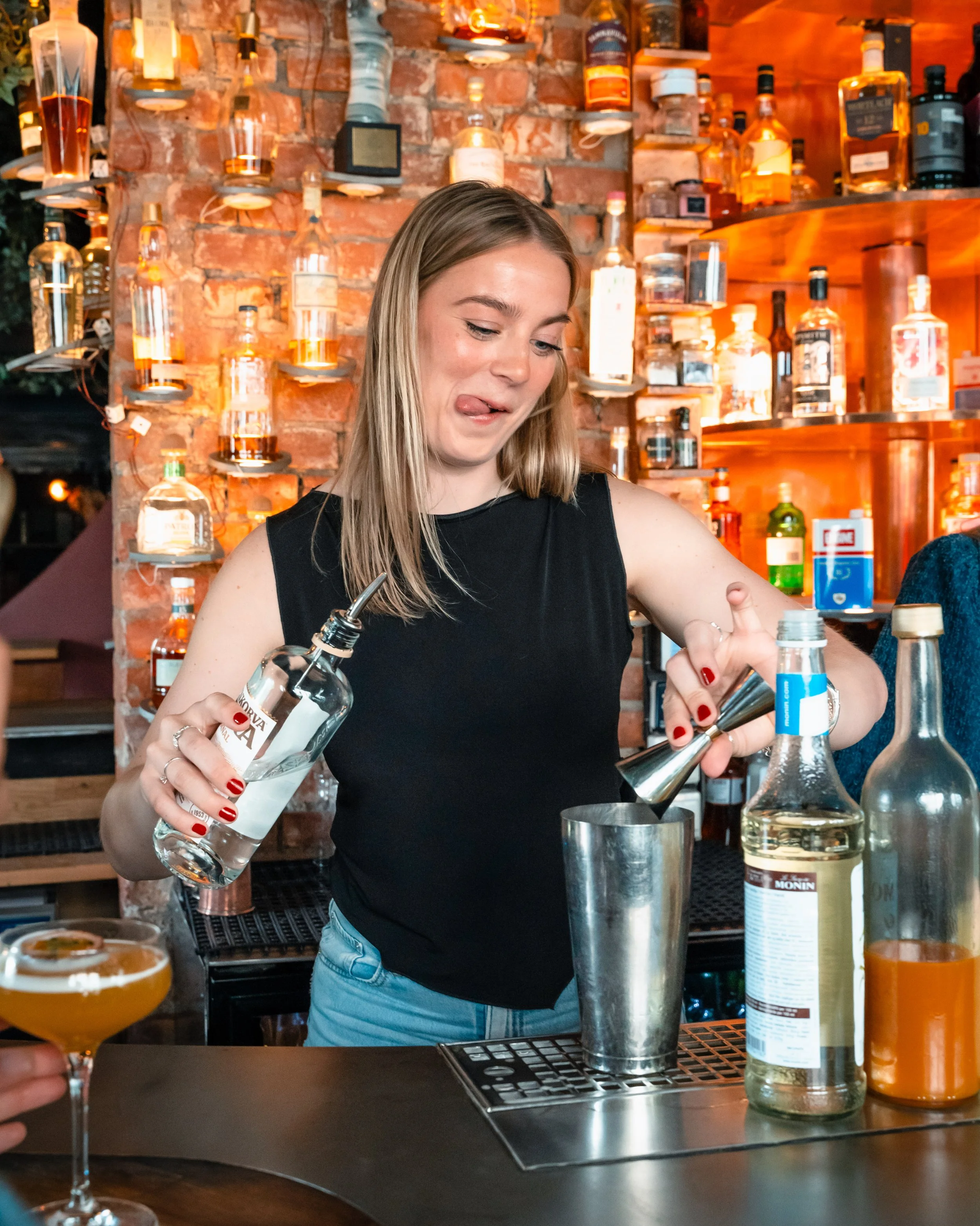 En kvinne som lager drinker bak baren med en murvegg og hyller med ulike flasker i bakgrunnen.