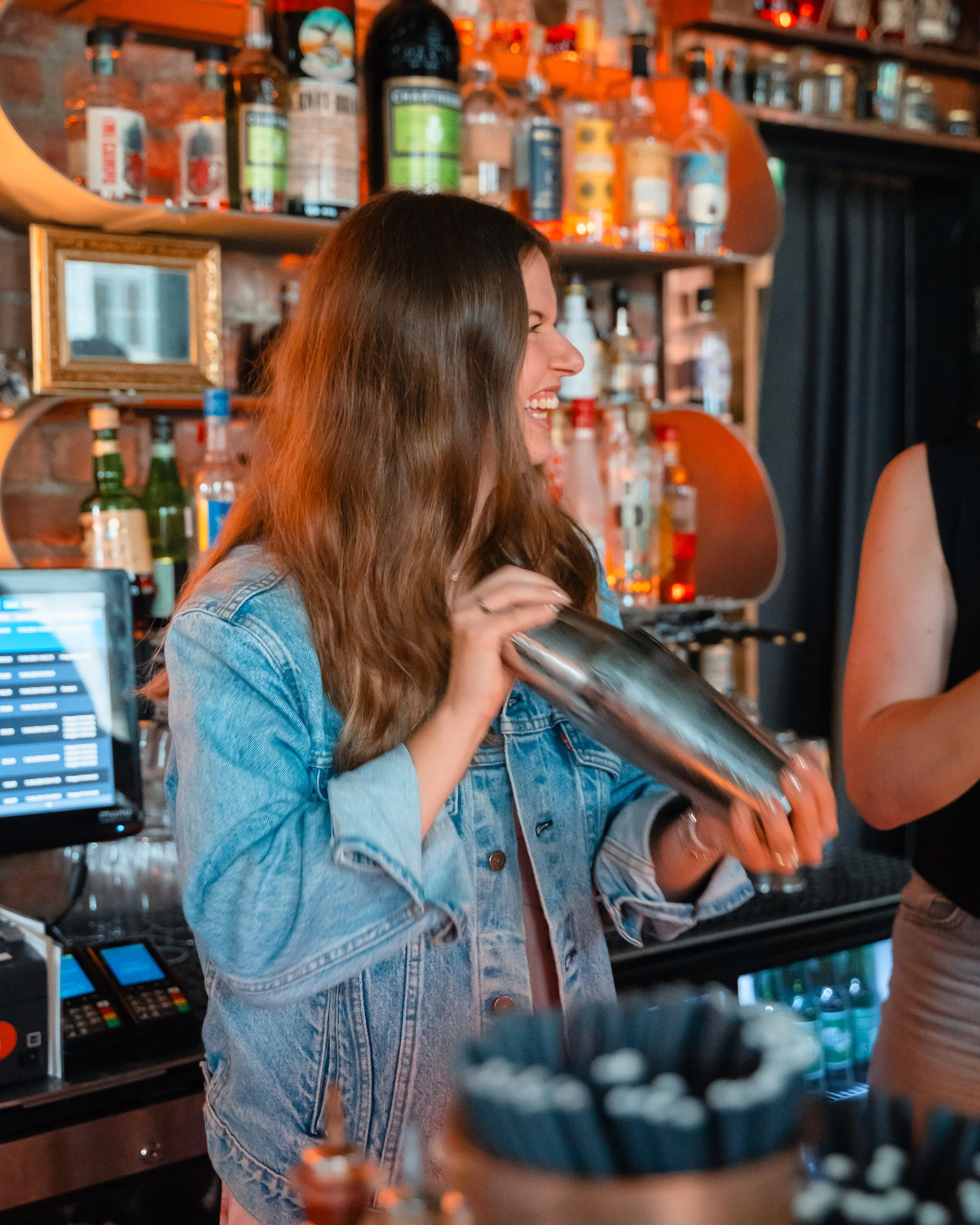 En kvinne med langt brunt hår ler og holder en metallic shaker i en bar, omgitt av alkoholflasker på hyller bak henne.