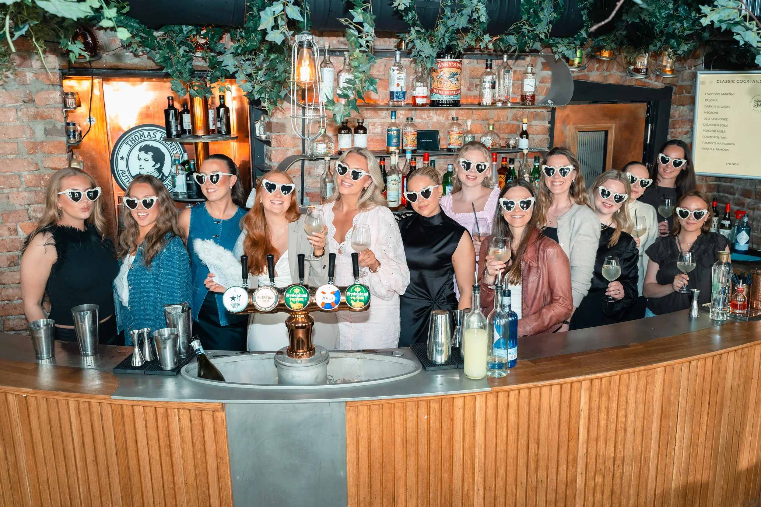 En gruppe kvinner med solbriller står bak baren i en bar eller restaurant, og de smiler og holder drinker. Det er en vegg med hyller fullt av flasker med alkohol på bakveggen.