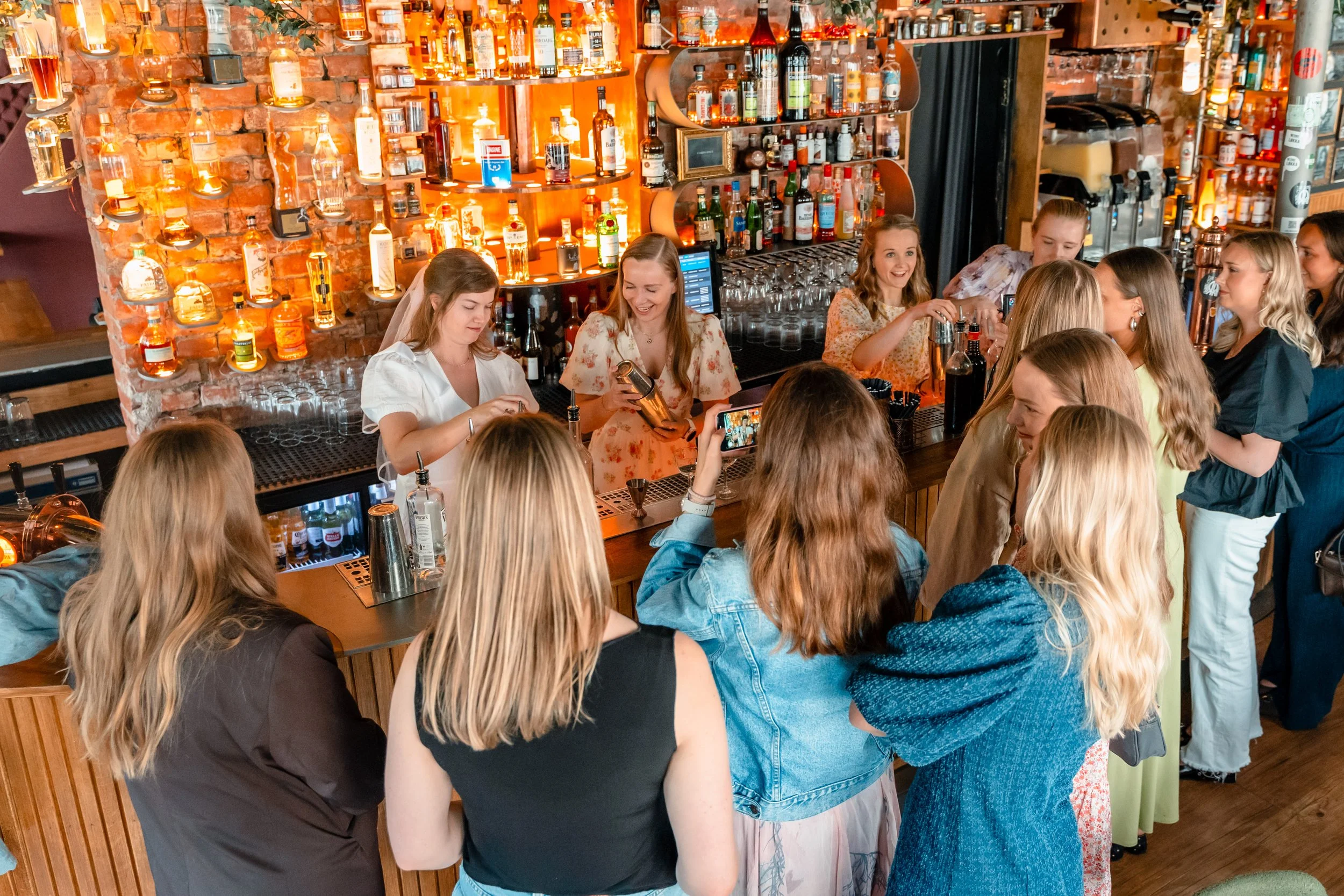En bar med mange mennesker, kvinner, som står i kø og bestiller drinker. Det er en rustikk bakvegg med hyller fulle av flasker med alkohol. Flere personer tar bilder av de som jobber bak baren.