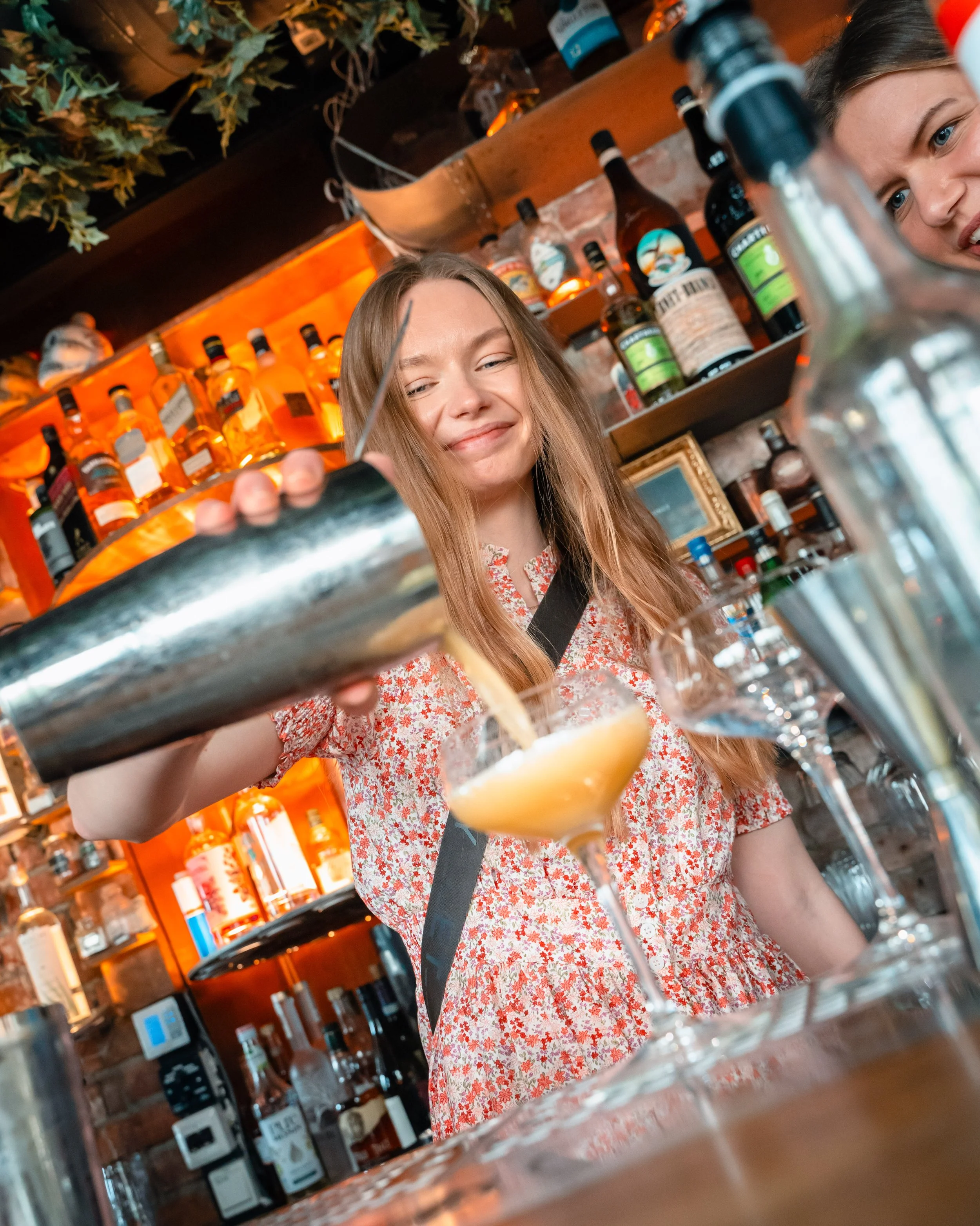 En kvinne med langt, lysbrunt hår og smilende ansikt som servitør bak baren, holder en cocktailglass med en gul drikke og sitronbåt. Baren har hyller med ulike alkoholflasker og dekorative elementer.