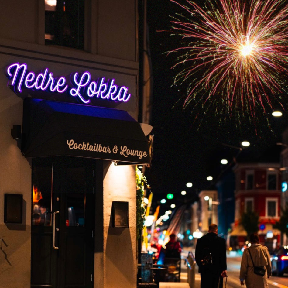 En gate om kvelden med mennesker som går, ferdig pyntet bygninger, en bygning med skilt som sier 'Nedre Lokka' og 'Cocktailbar & Lounge', og fyrverkeri på himmelen.