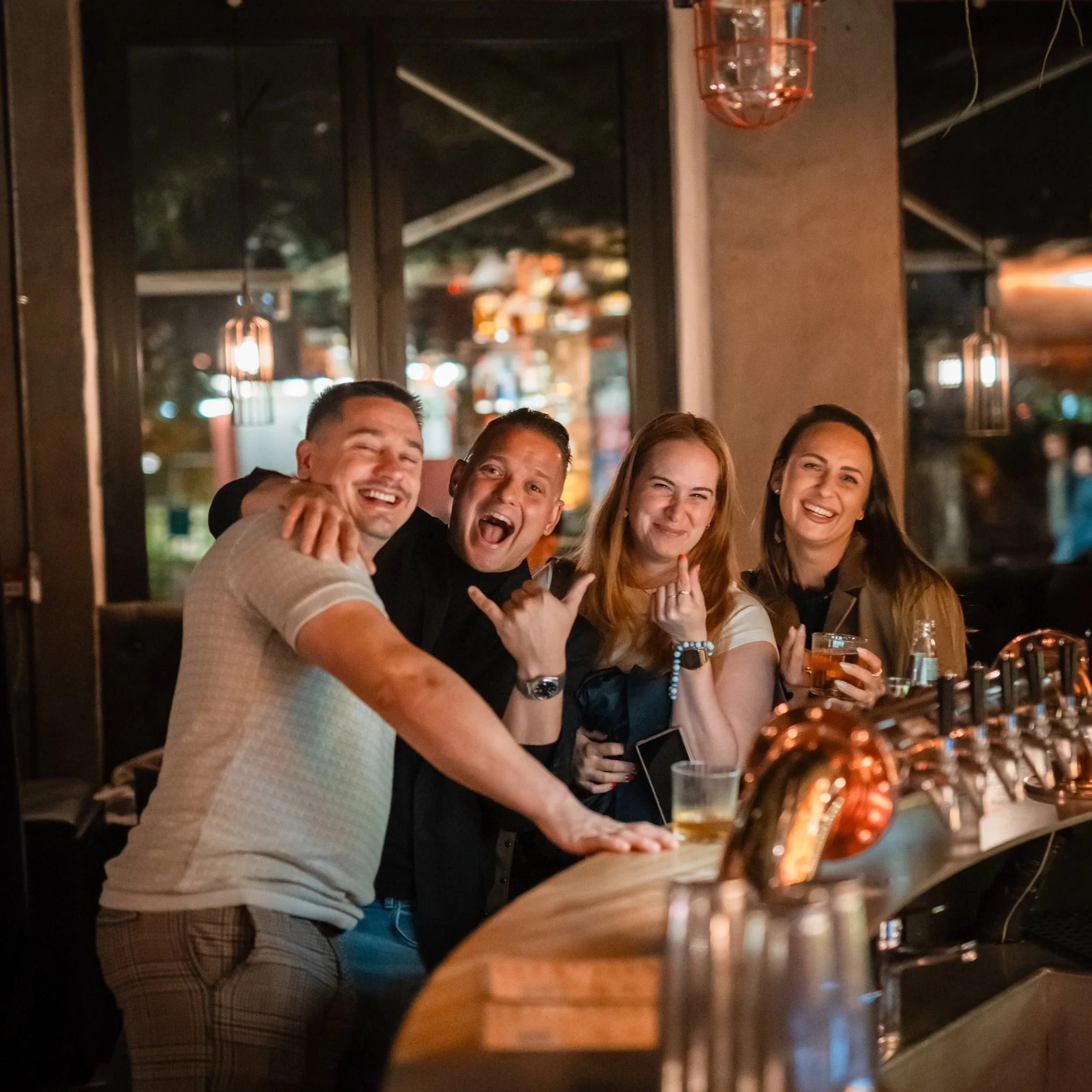 Bildet viser en gruppe mennesker som smiler og koser seg, sittende på en bar eller restaurant. Bakgrunn med store vinduer og blurrende lys.
