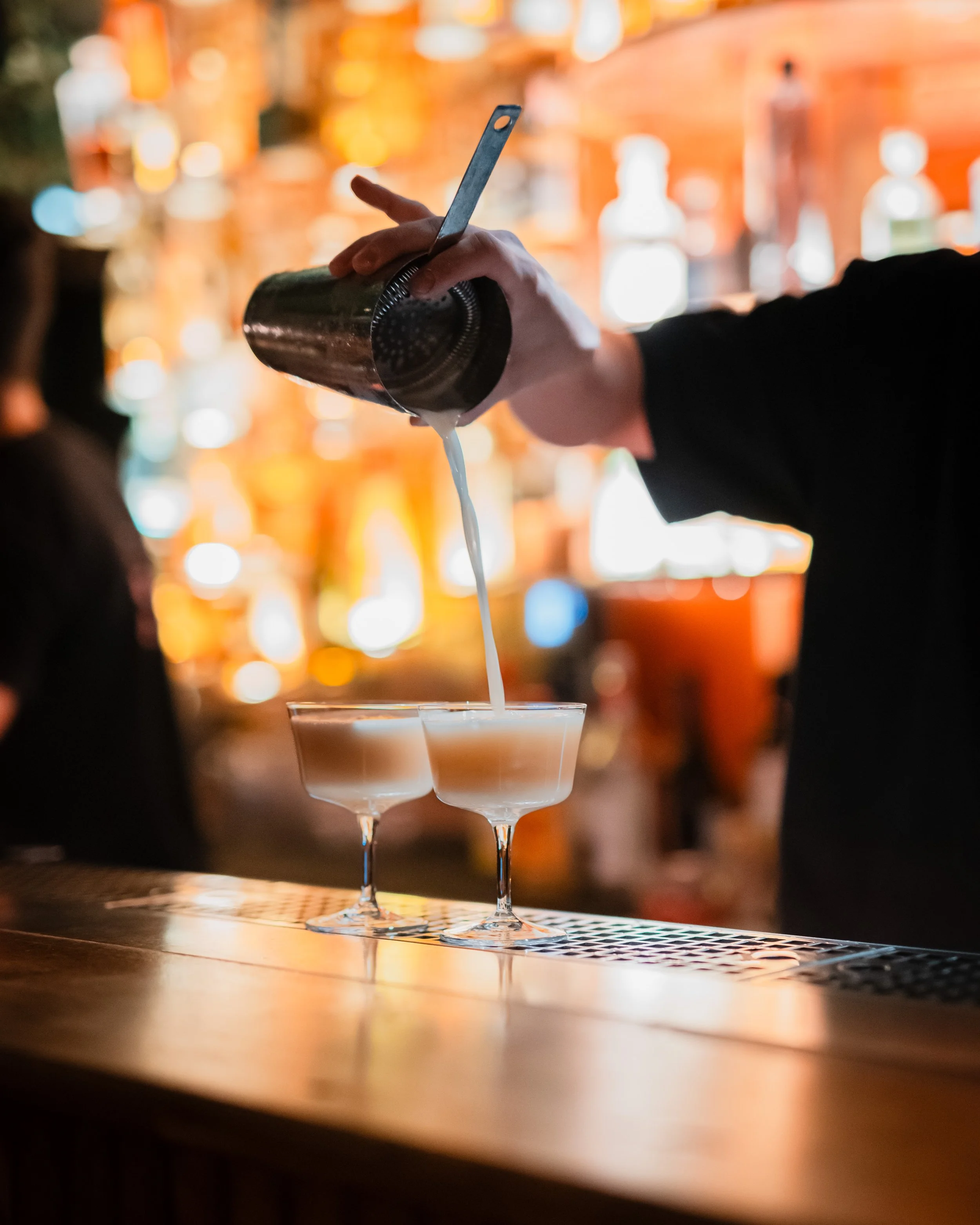 Bilde av en person som heller en hvitflytende drink fra en metallshaker i et cocktailglass på en bar med varmt, dimmet lys i bakgrunnen.
