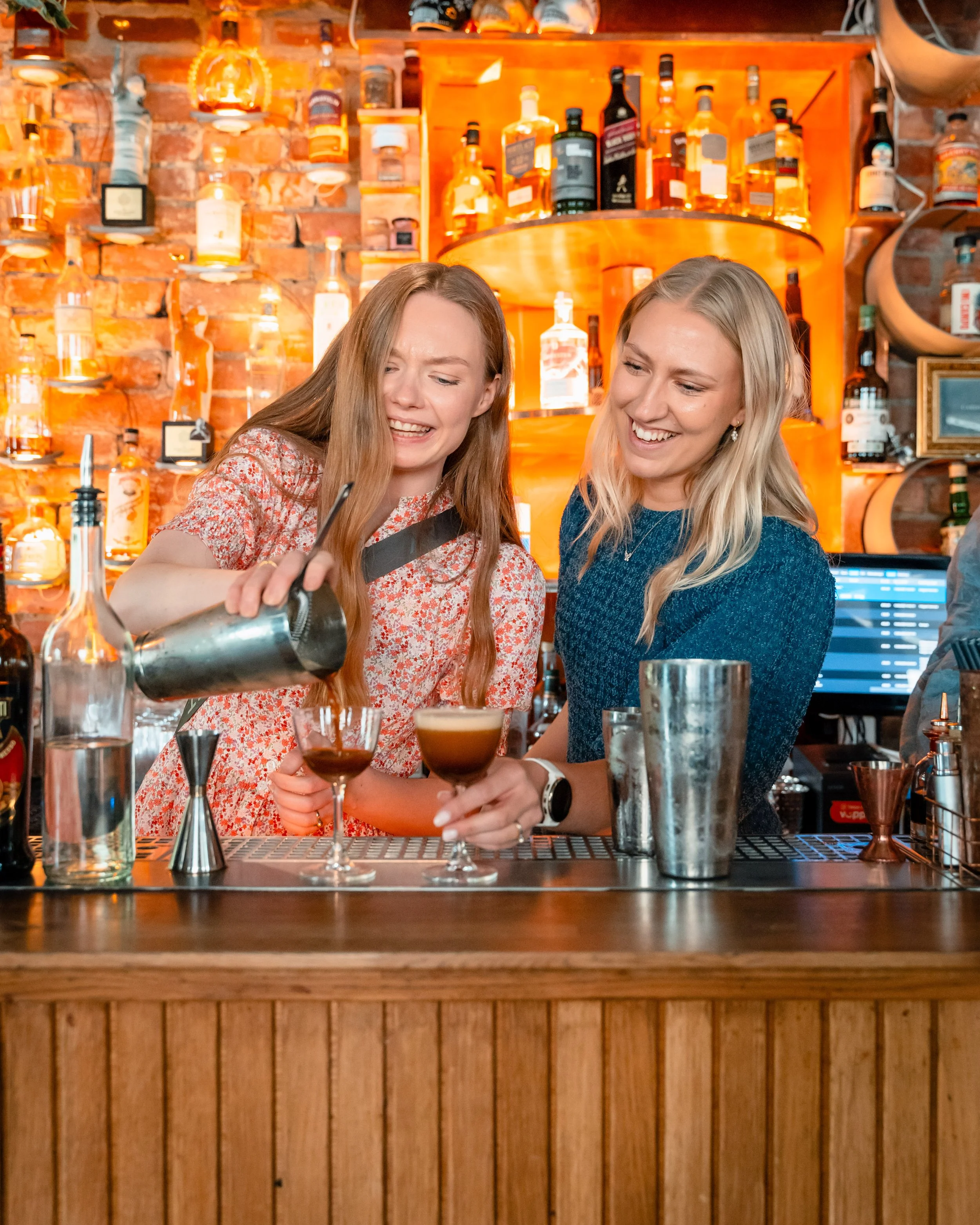 To kvinner som smiler og snakker på en bar, der den ene heller opp en cocktail. Baren har en bakvegg med mange flasker av forskjellige typer alkohol, og det er tre mennesker i bildet.