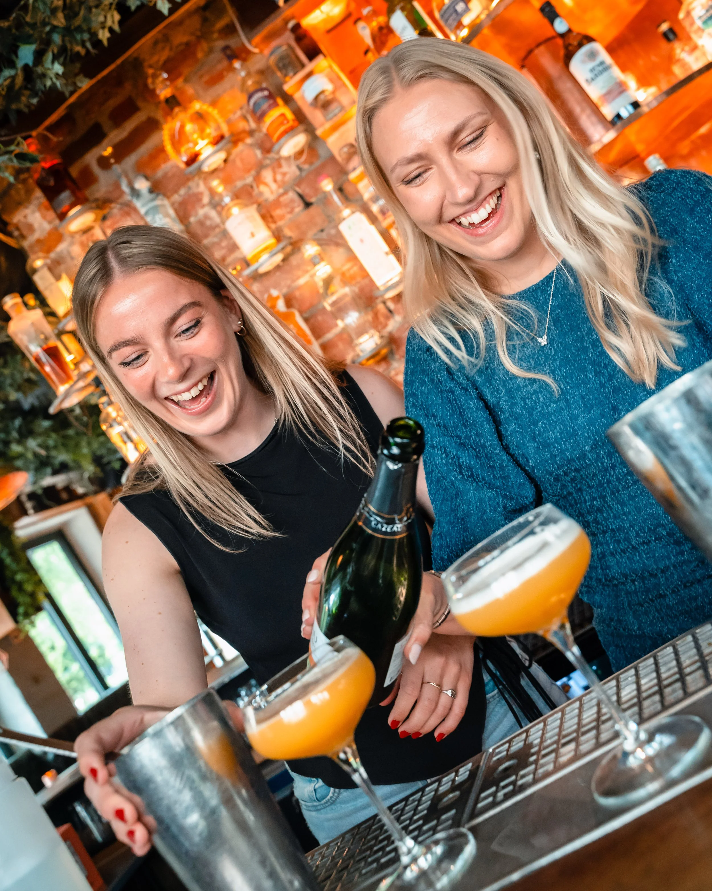 To kvinner smiler mens de lager hvitvin i en bar eller restaurant, med en bar i bakgrunnen og drikke glass foran dem.