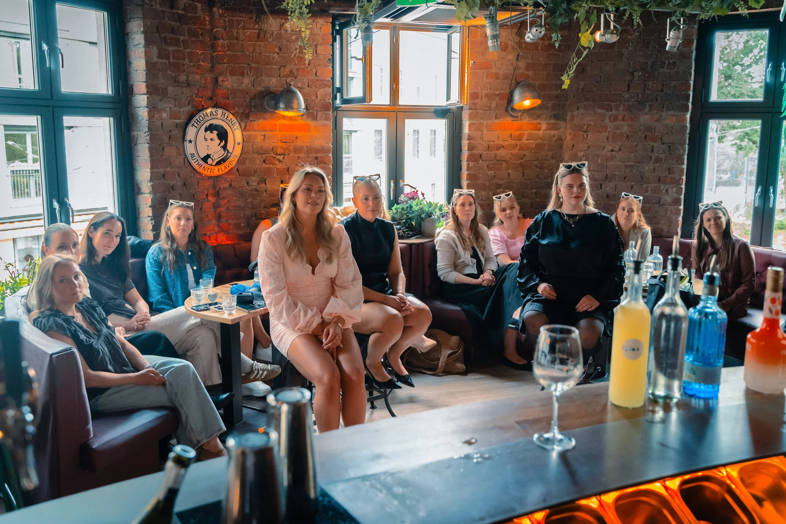 En gruppe mennesker sitter rundt et langbord i en restaurant eller kafé med røde murvegger og store vinduer. Det ser ut til å være en samling eller møte, og flere av personene har solbriller. Det er ulike drikker og en flaske på bordet, og i bakgrunn