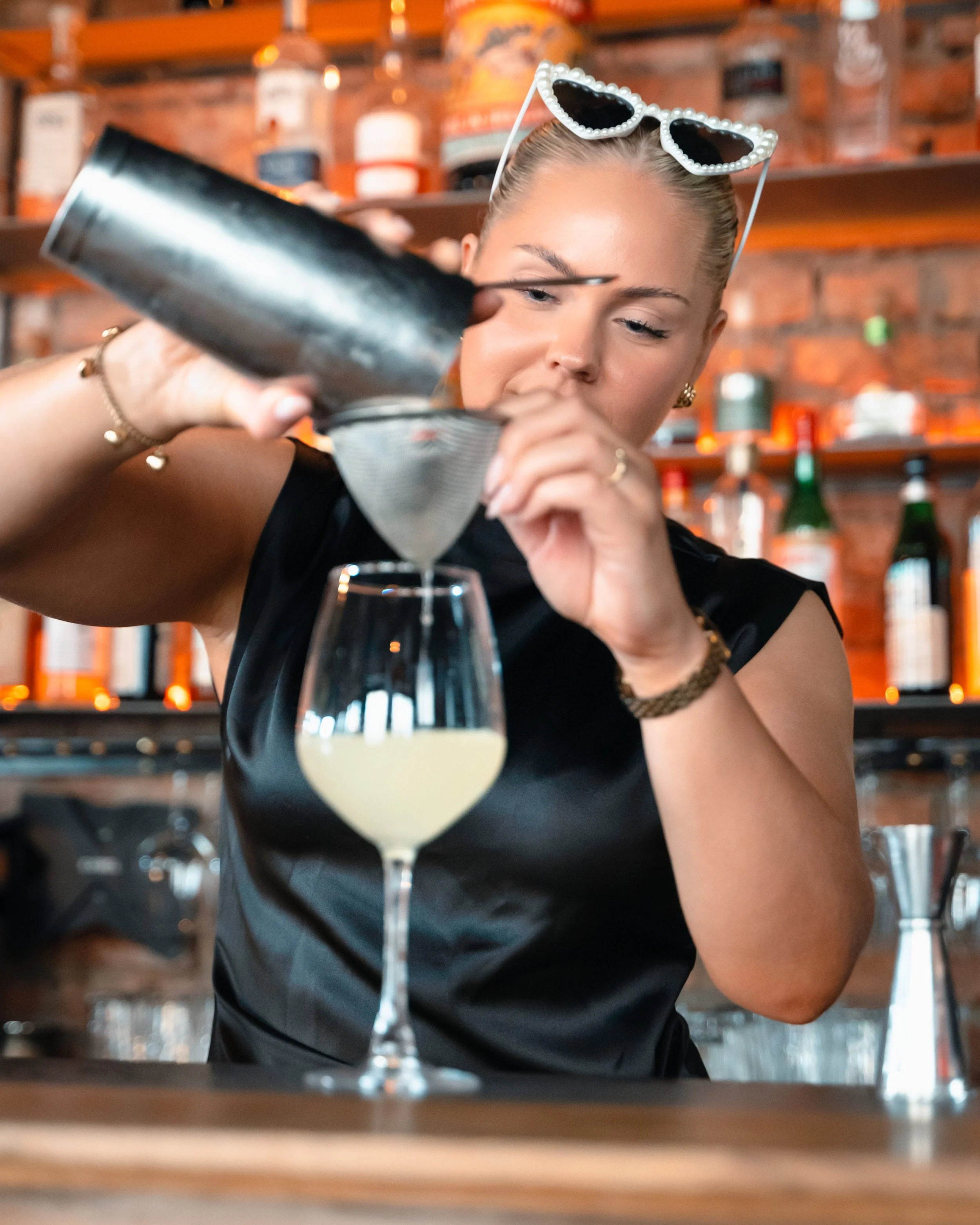 En kvinne med solbriller med perleinnfatning, som lager en cocktail på en bar med en rustikk, eksponert murvegg i bakgrunnen.