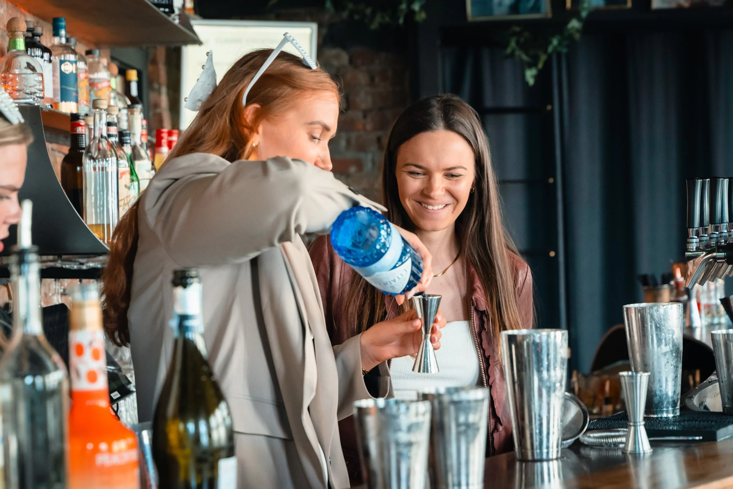 To kvinner står bak baren, en heler is og drinker. Den ene av dem heler en blå drink i et målebeger. Det er flere flasker og drinkutstyr på bardisken.