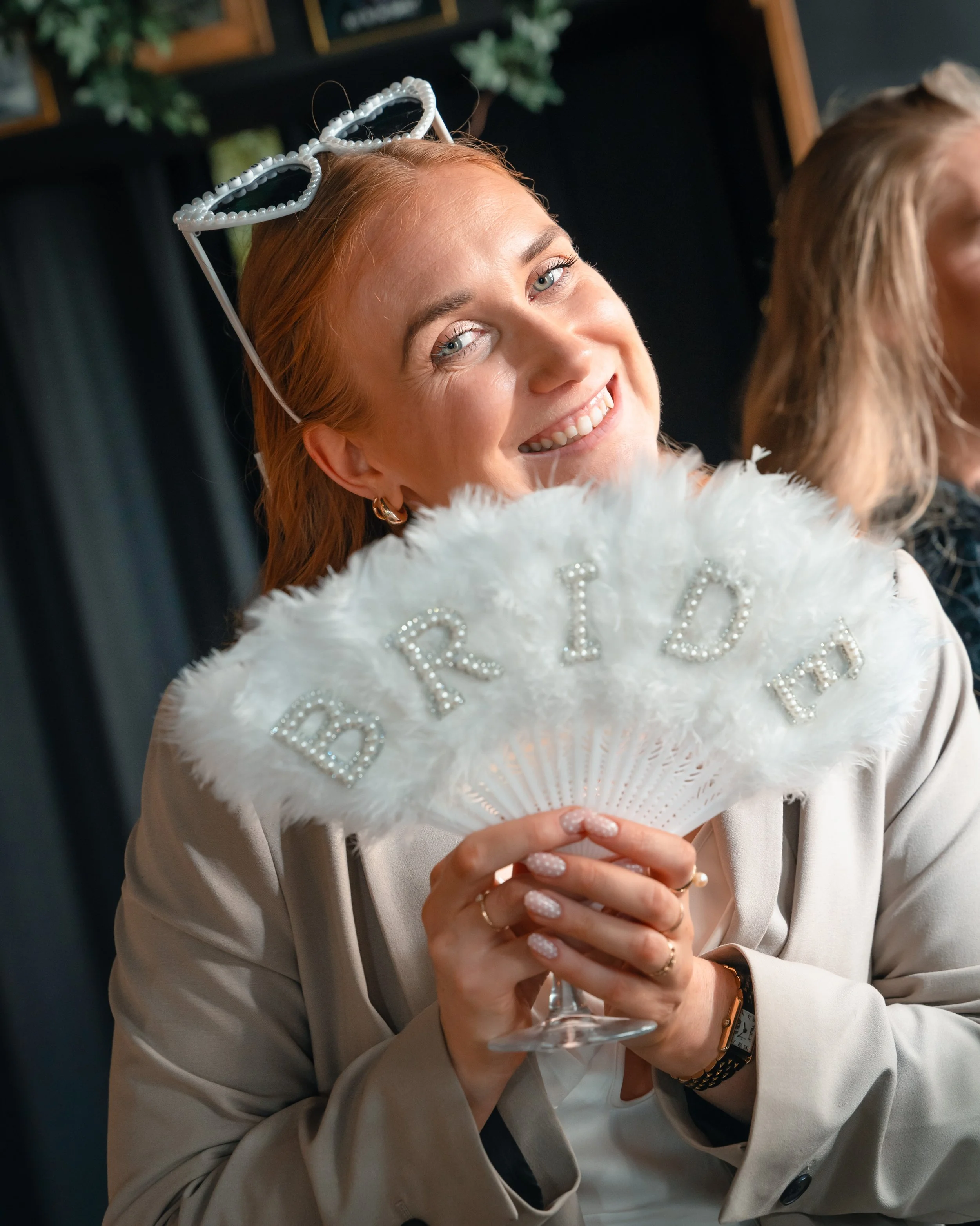 En kvinne med rødt hår, holder en glass med champagne og smiler. Hun har på seg en beige blazer, har peacetato og en armbåndsur. På hodet har hun en hjerteformet briller laget av perler.