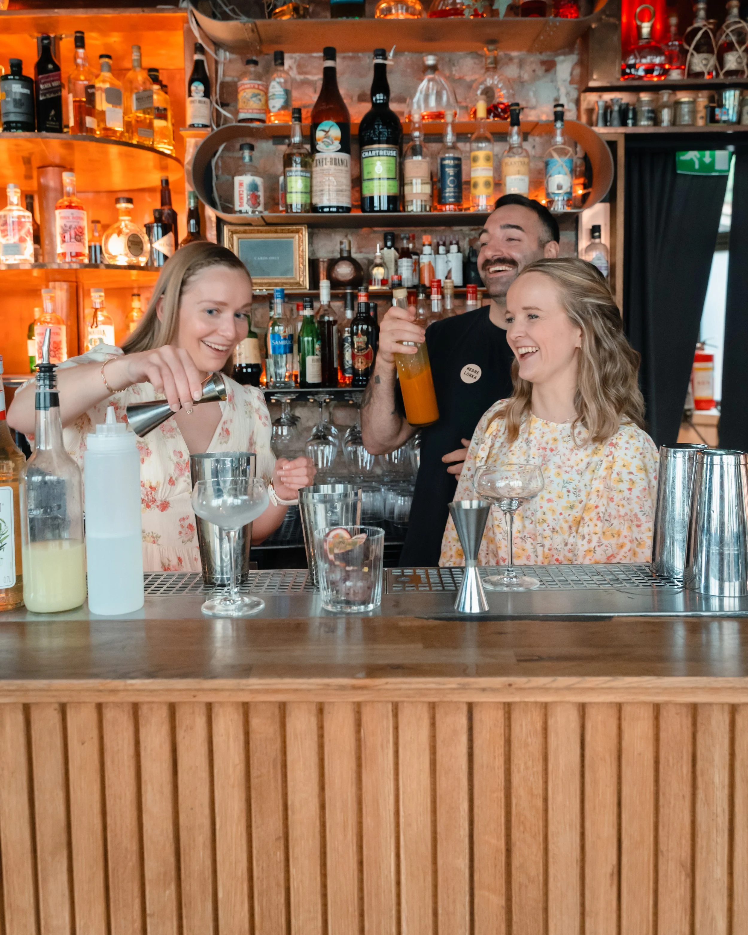 Bart med ulike cocktail- og alkoholglass, og tre personer bak baren smilende og føre samtaler i en bar.