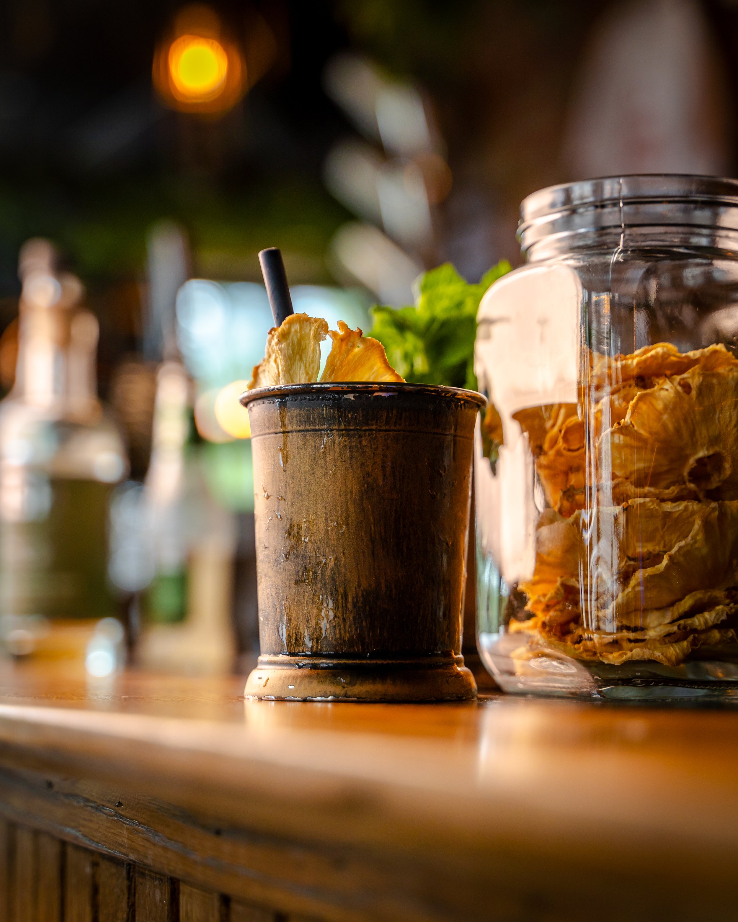 Et glass med fruktchips og friske urter, en glasskrukke med flere fruktchips, satt på en trebord i et rolig, utendørs miljø med bokeh-lys i bakgrunnen.