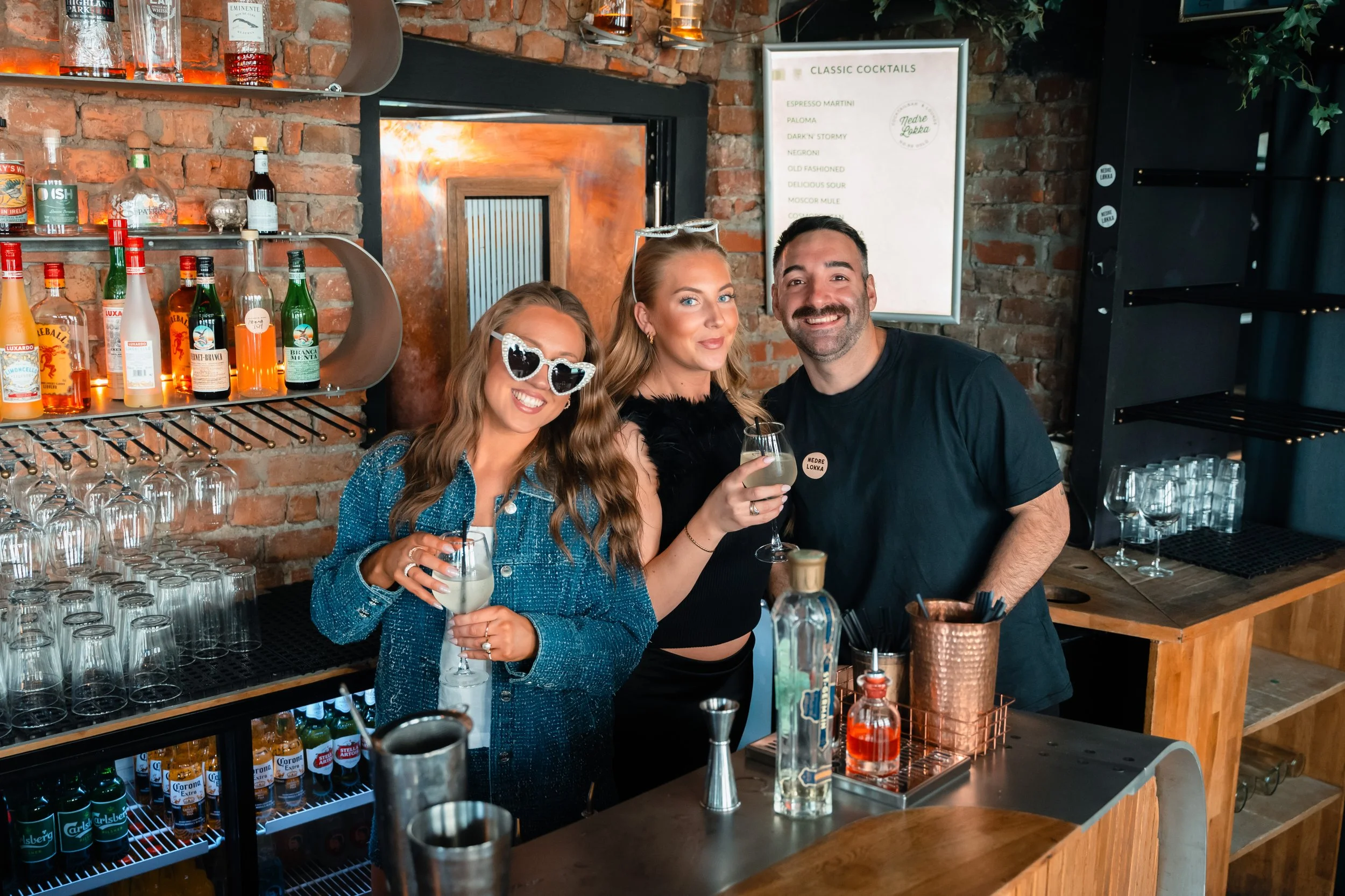 Tre personer som smiler og holder drinker i en bar. Bak en hylle med ulike flasker alkohol og et utvalg av glass. En meny med cocktailnavn til høyre.