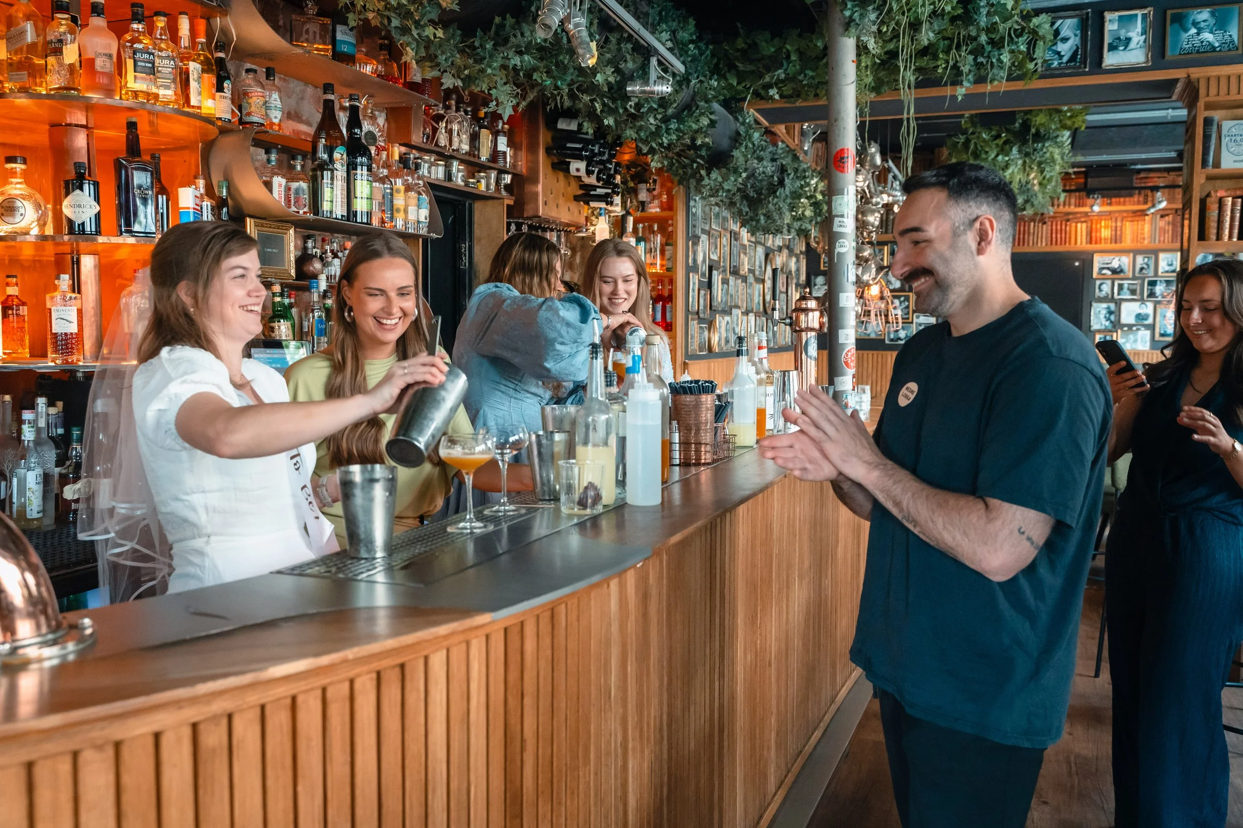 En gruppe unge mennesker som ler og snakker på en bar, med drikker og bartendere som serverer i en innendørs bar med pubelementer og grønne planter.