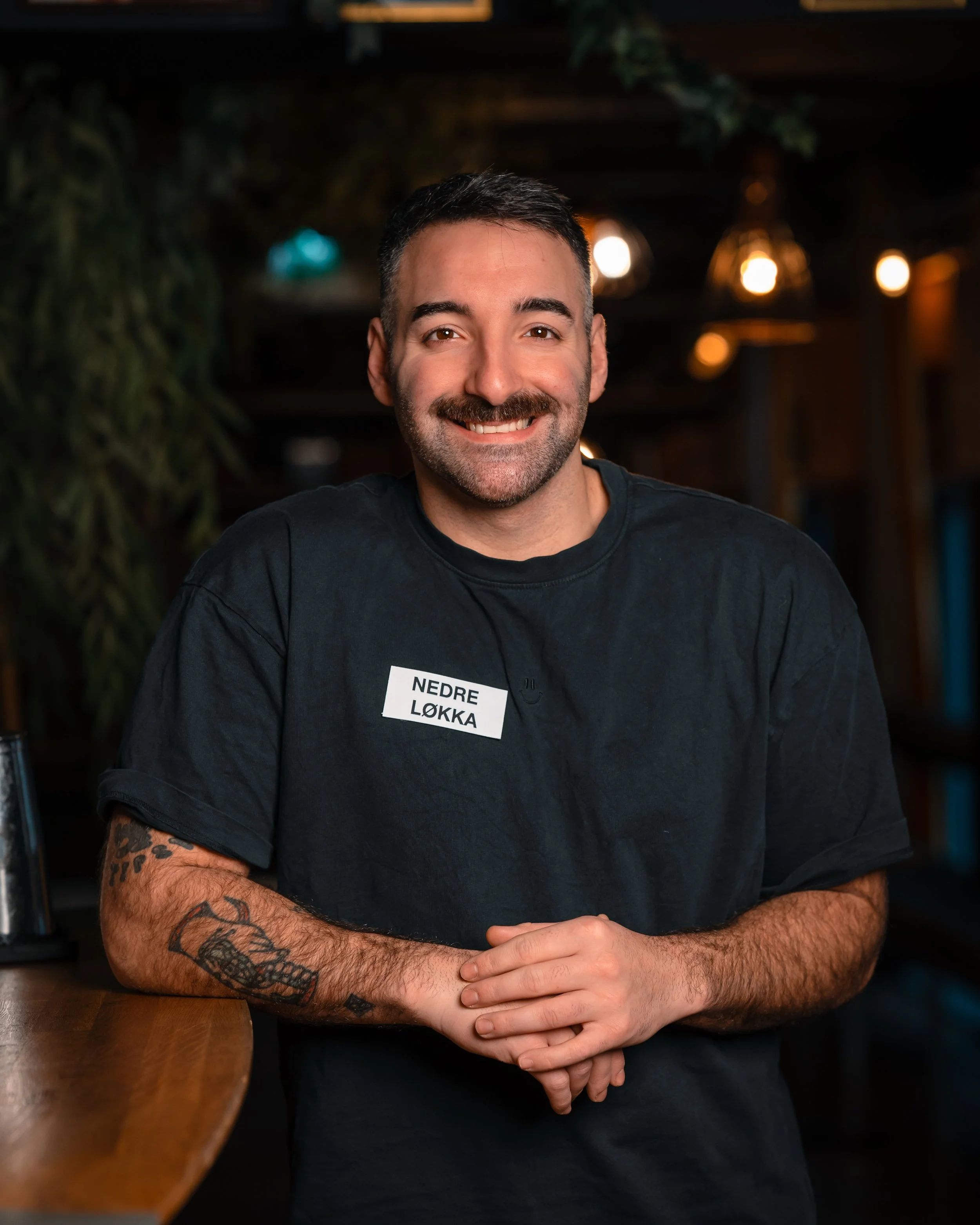 Et nærbilde av en smilende mann med kort svart hår, skjegg og tatoveringer på armen, sittende ved et tavlebord i en bar eller restaurant med varmt belysning. Han har på seg en svart t-skjorte med hvitt tekst.