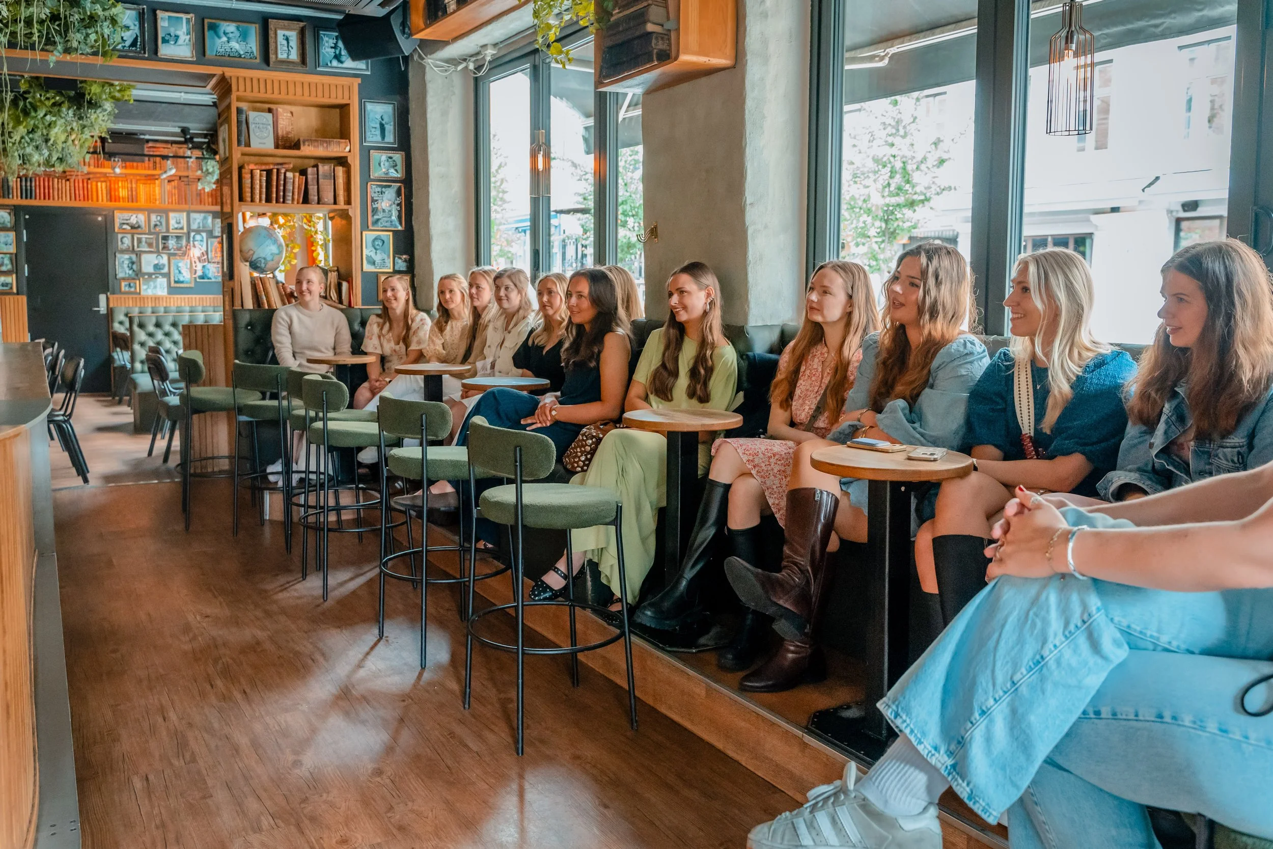 En gruppe med kvinner sitter i en kafé og ser mot venstre, mens de smiler og ser oppmerksomt. Det store glassdøret i bakgrunnen slipper inn mye naturlig lys, og veggen er dekorert med bilder og bøker.