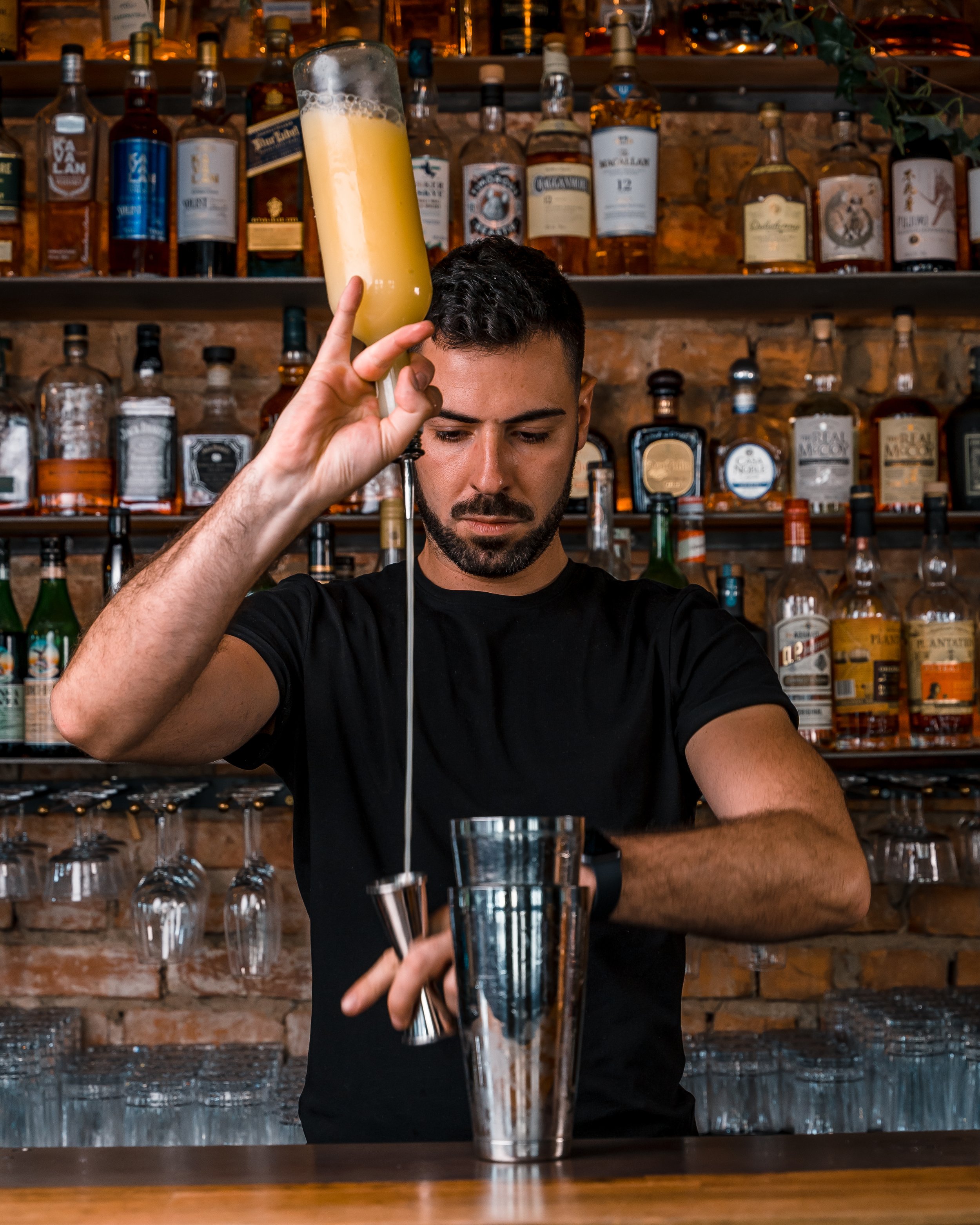 En mann som lager en cocktail bak baren, med flasker med alkohol på hyllen i bakgrunnen.