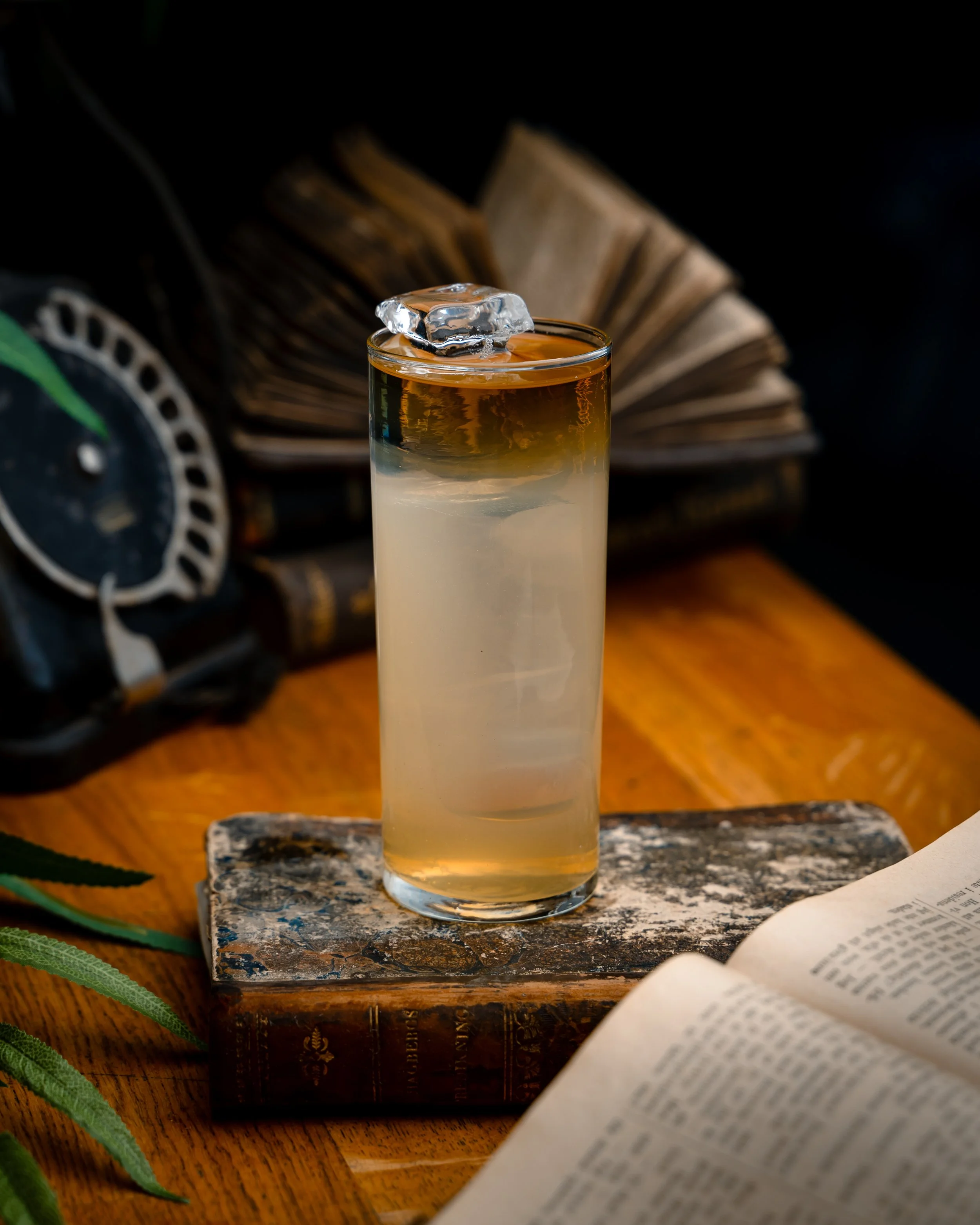 Et glass med en to-farget cocktail, toppet med et isstykke, plassert på et gammelt, rustent treskjold med en åpen bok og et blad i forgrunnen, og en bok i bakgrunnen.