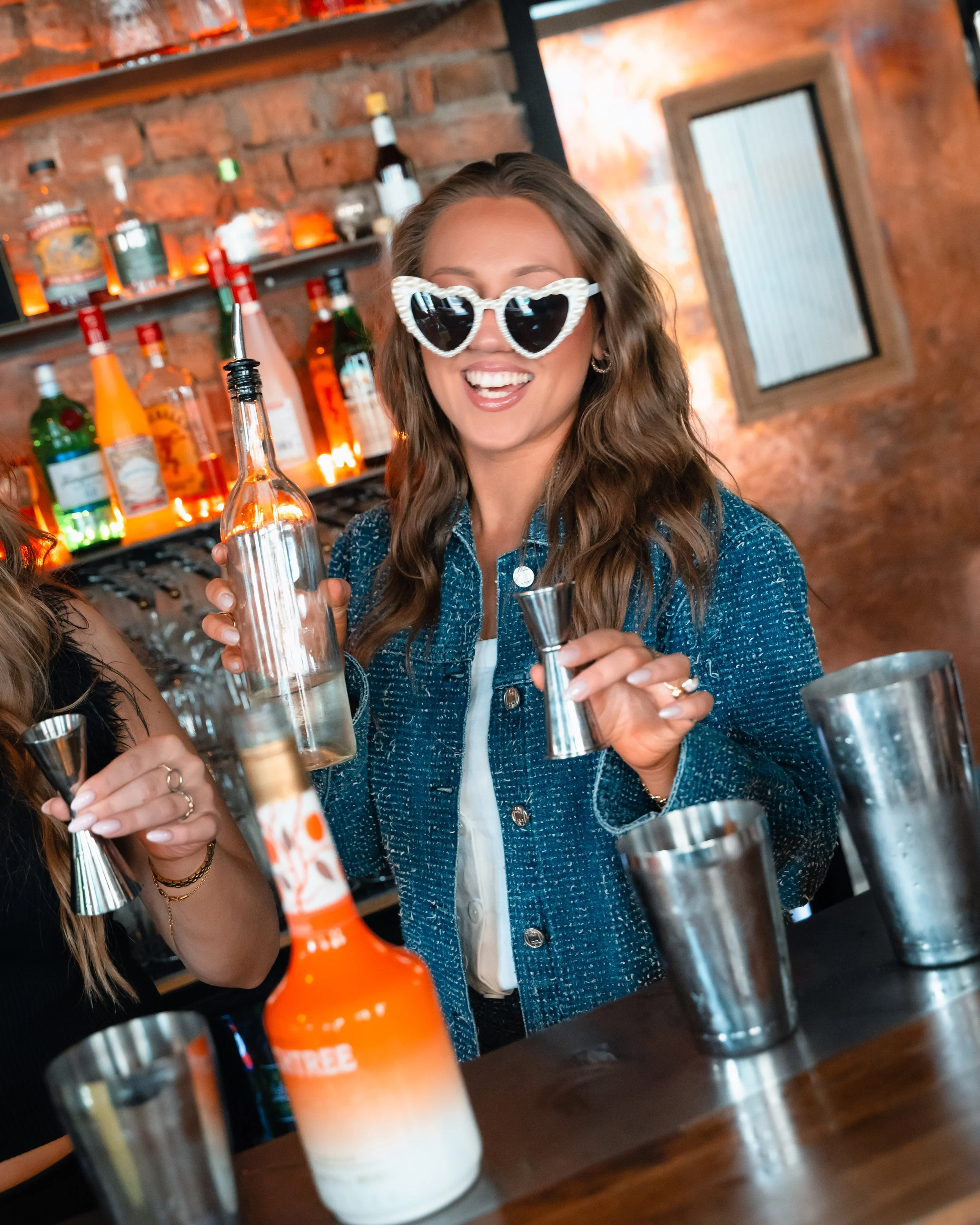 En jente med krøllete brunt hår som smiler med store, hvite hjertelignende solbriller i en bar, holdende en tom klar brusbottle. Røffe murvegger og barhyller med ulike alkoholflasker i bakgrunnen.