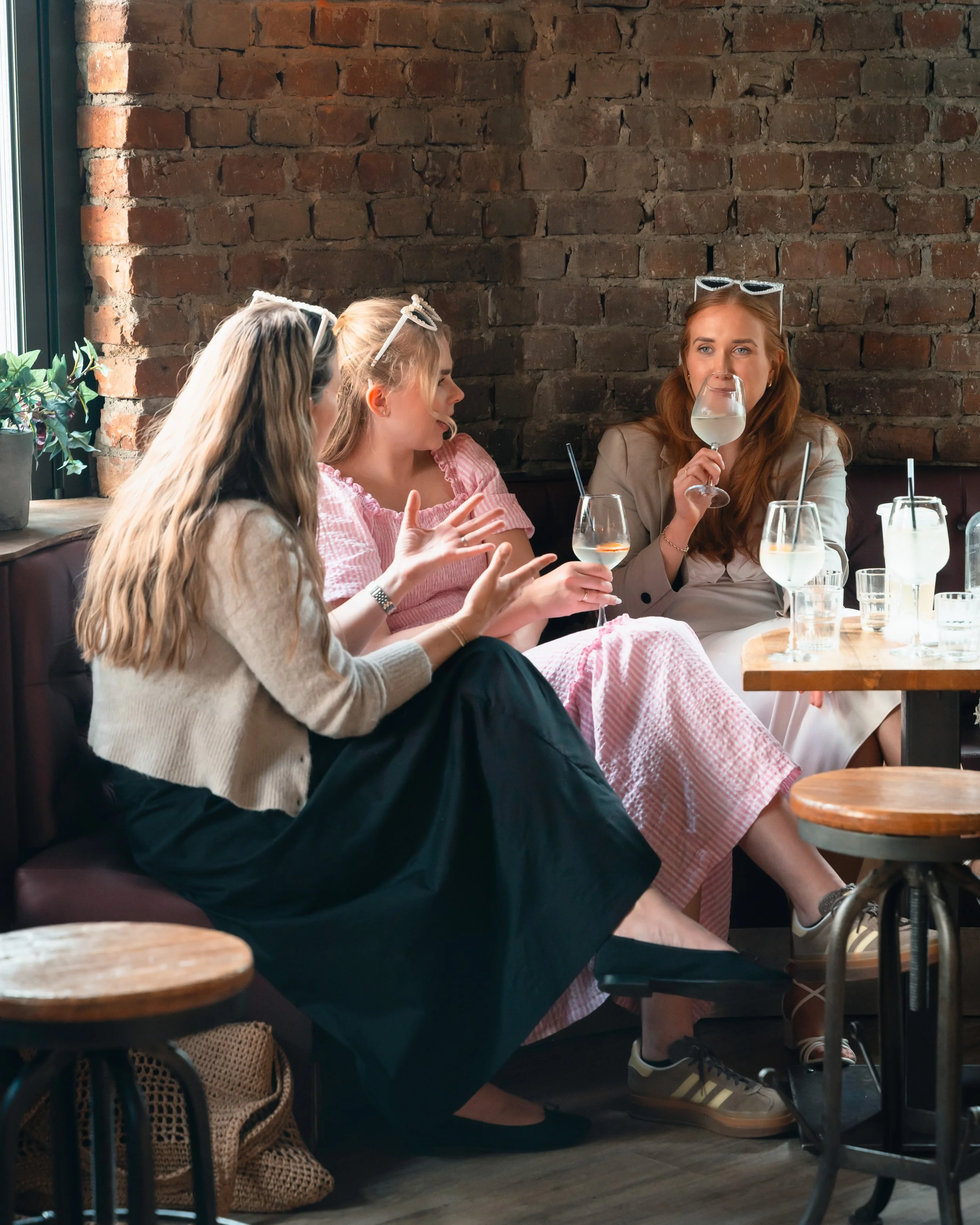 Tre kvinner sitter på en kafé, drikker hvitvin og har samtale. Bakgrunnen er en murvegg. Det er noe grønt planteliv på venstre side.