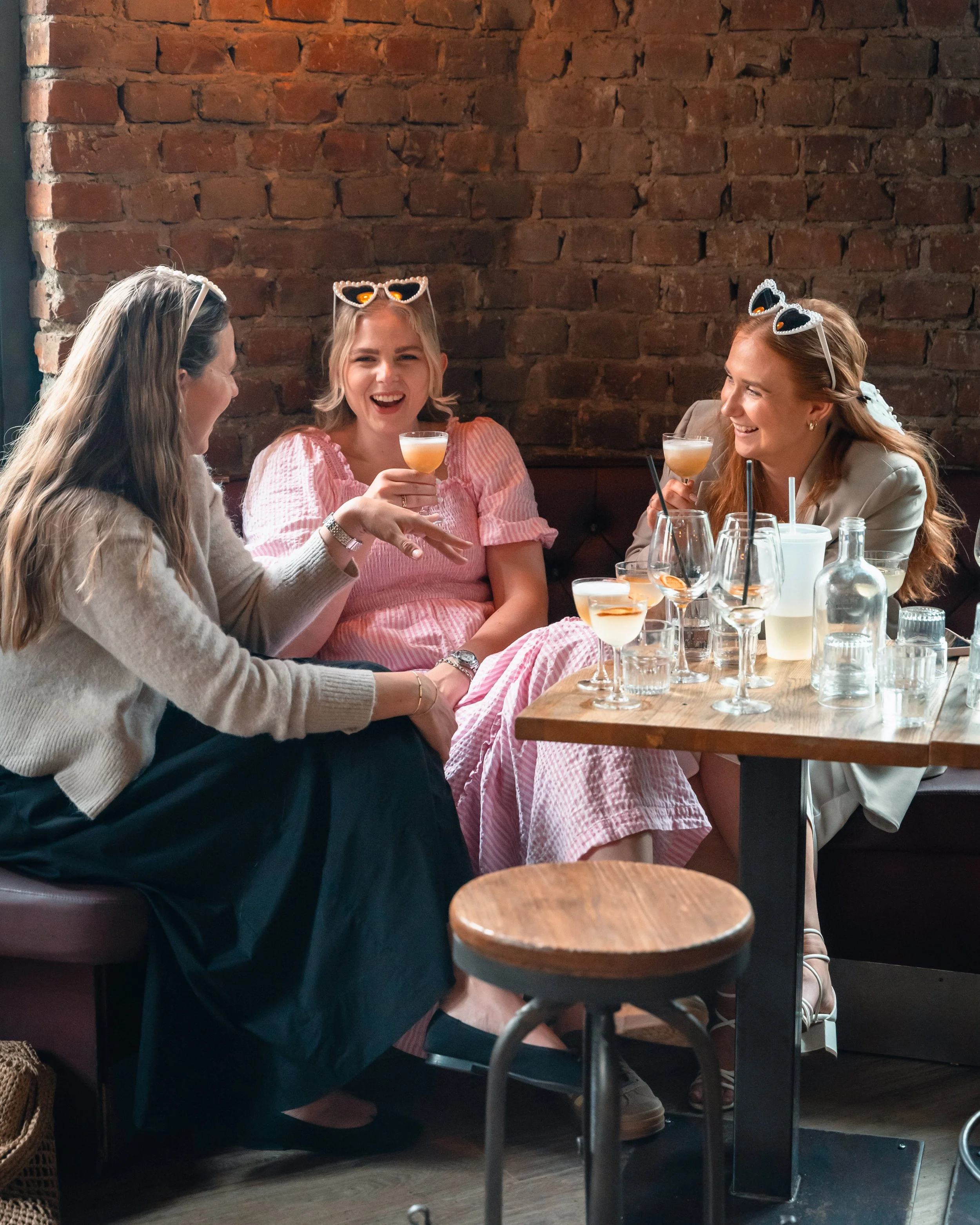 Tre kvinner sitter rundt et bord på en restaurant, ler og snakker. De har drinker i hånden og har på seg solbriller. Bakgrunnen har en ubrukt mursteinsvegg.