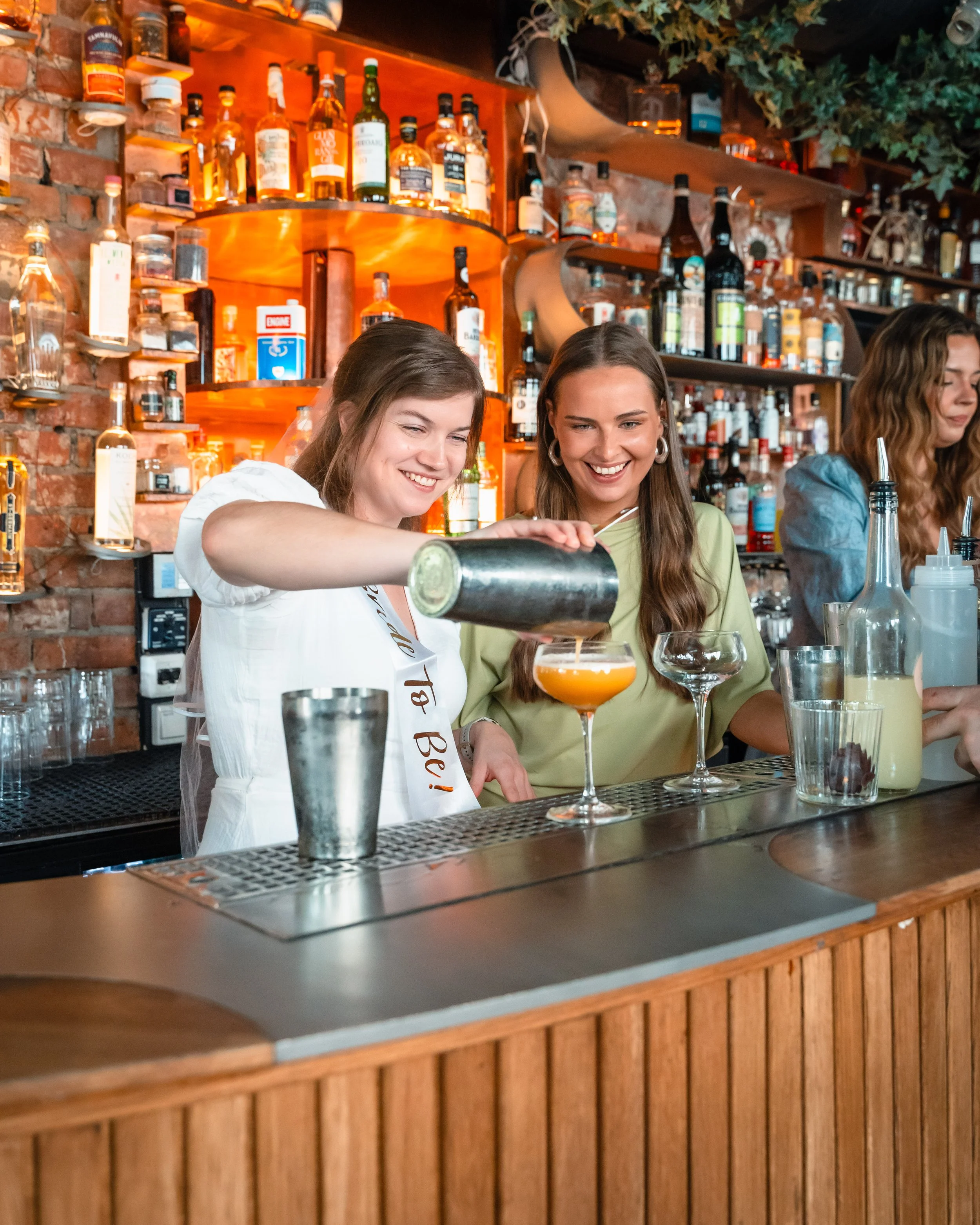 To kvinner står bak baren og lager drinker, med en bakvegg av godt utvalg av brennevin. Den har tre kvinner, en med hvit skjorte og en i grønn topp, og en annen med blå skjorte. Det er en rustikk bar med murvegger og trehyller.