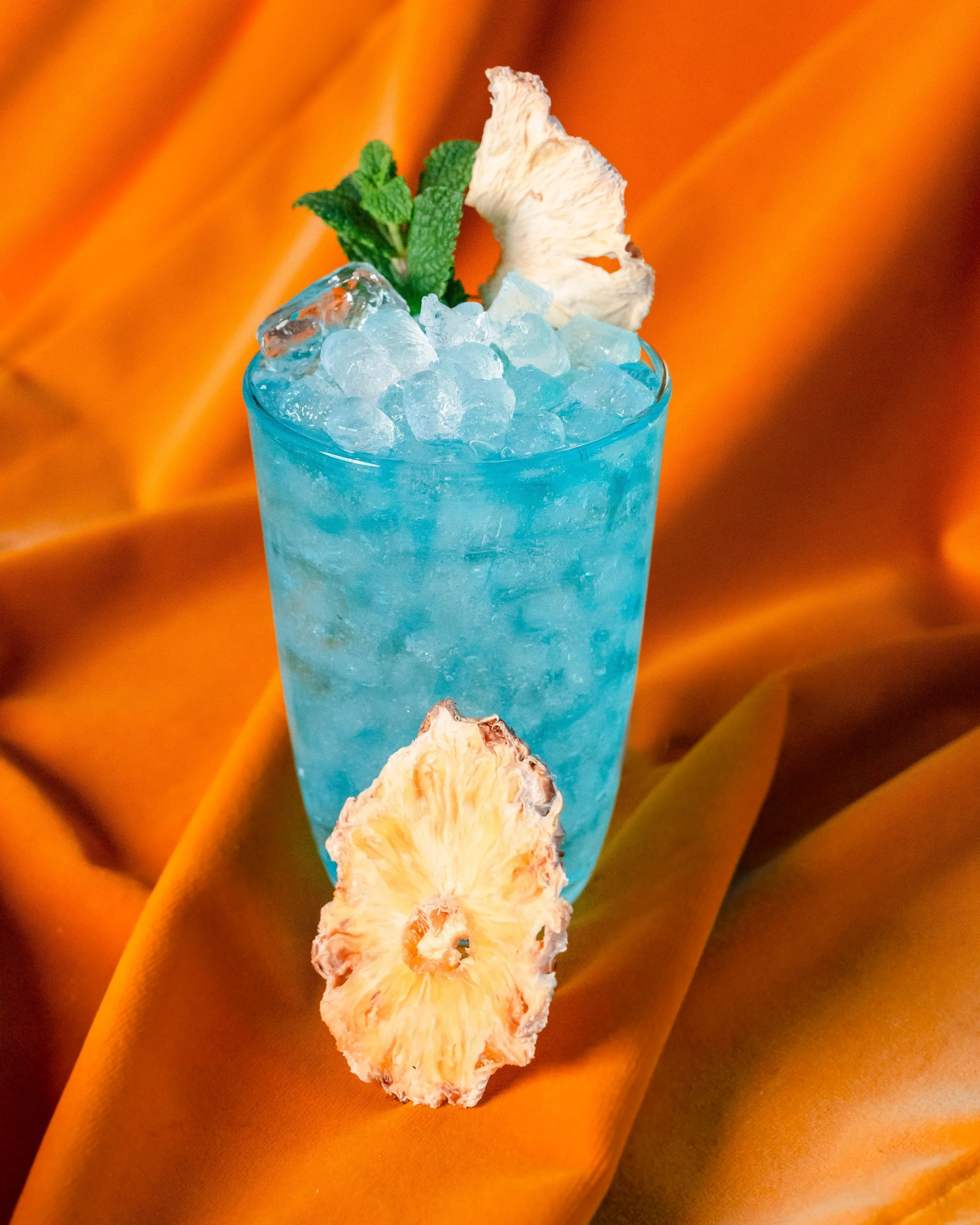 Et glass med blå drink, pyntet med en myntebunt, tørket ananas og enkelte isbiter. Bakgrunnen er oransje og gul satengstoff.