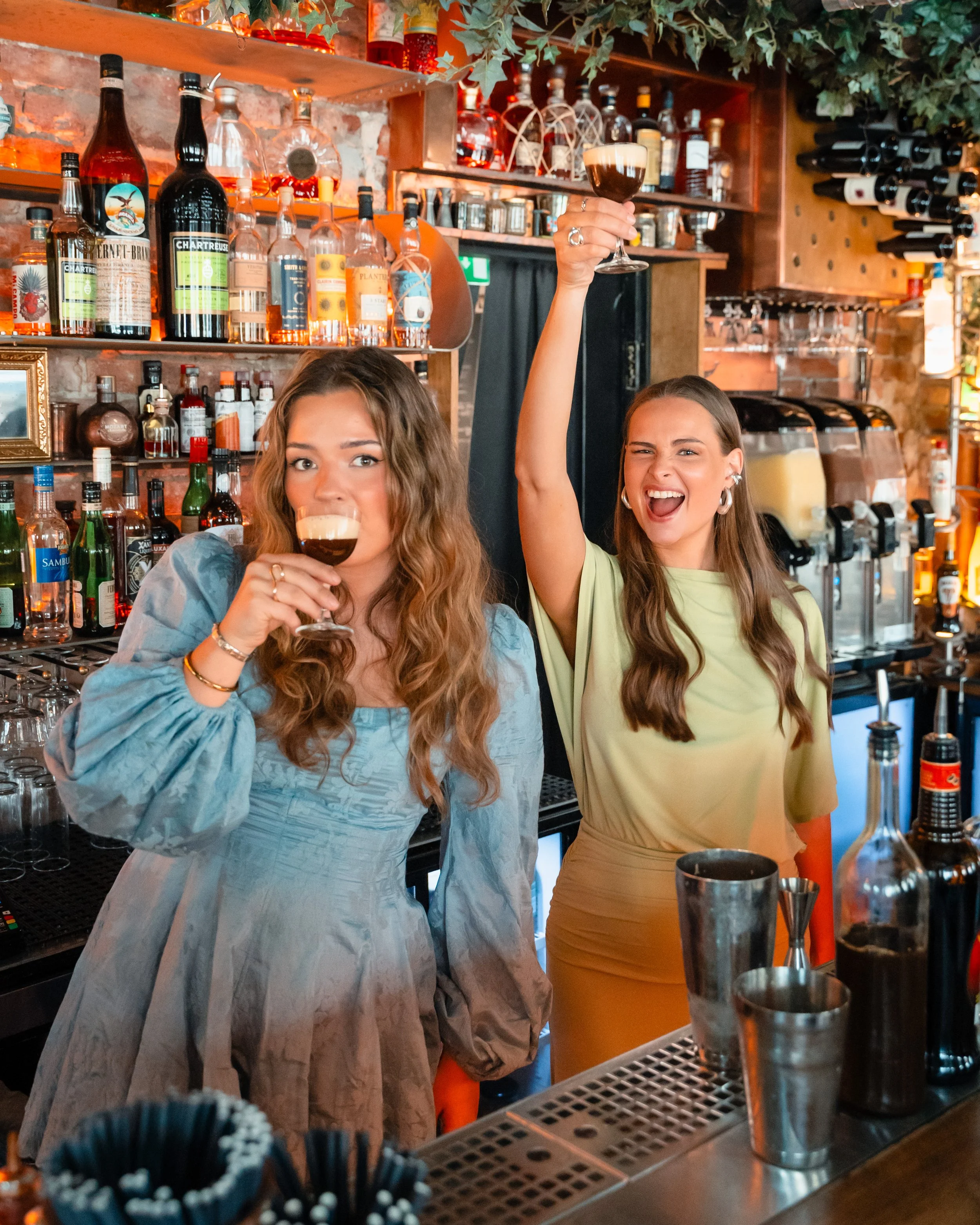 To kvinner foran et bartender-område, den ene holder et glass med mørk drink og det andre drikker av et glass, i en bar med mange flasker og utstyr.