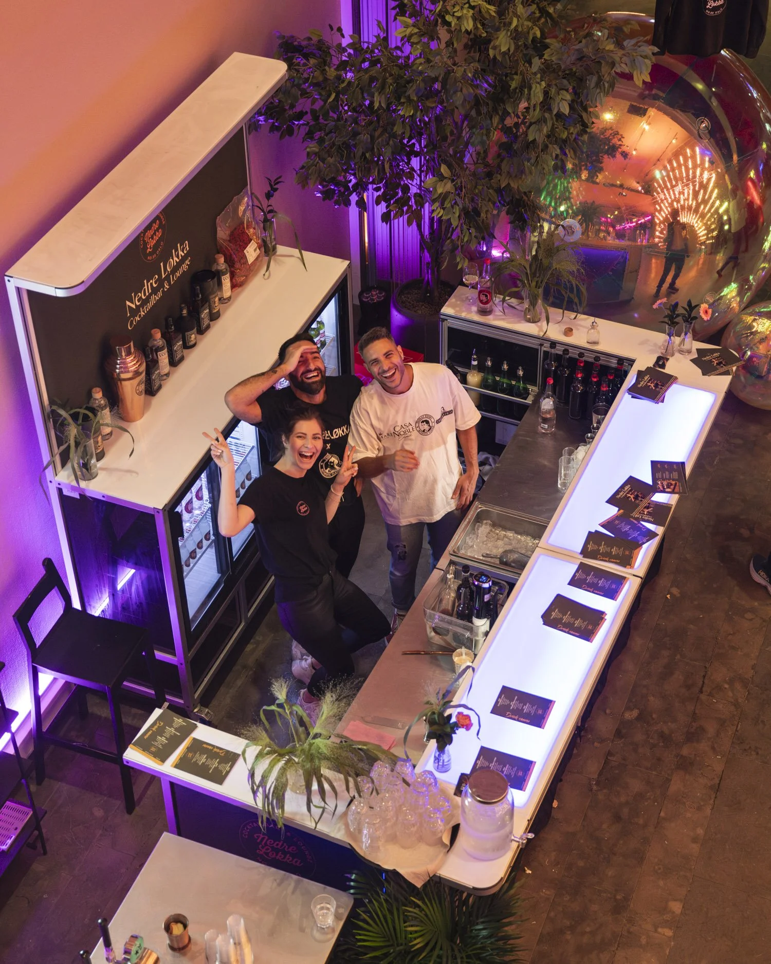 Bildet viser en bar på en nattklubb med tre personer som smiler og poserer for kameraet. Det er et opplyst barområde med planter og refleksjoner av lys i en stor rund dekorasjon på veggen. Det er også lysende menyelementer på bardisken.