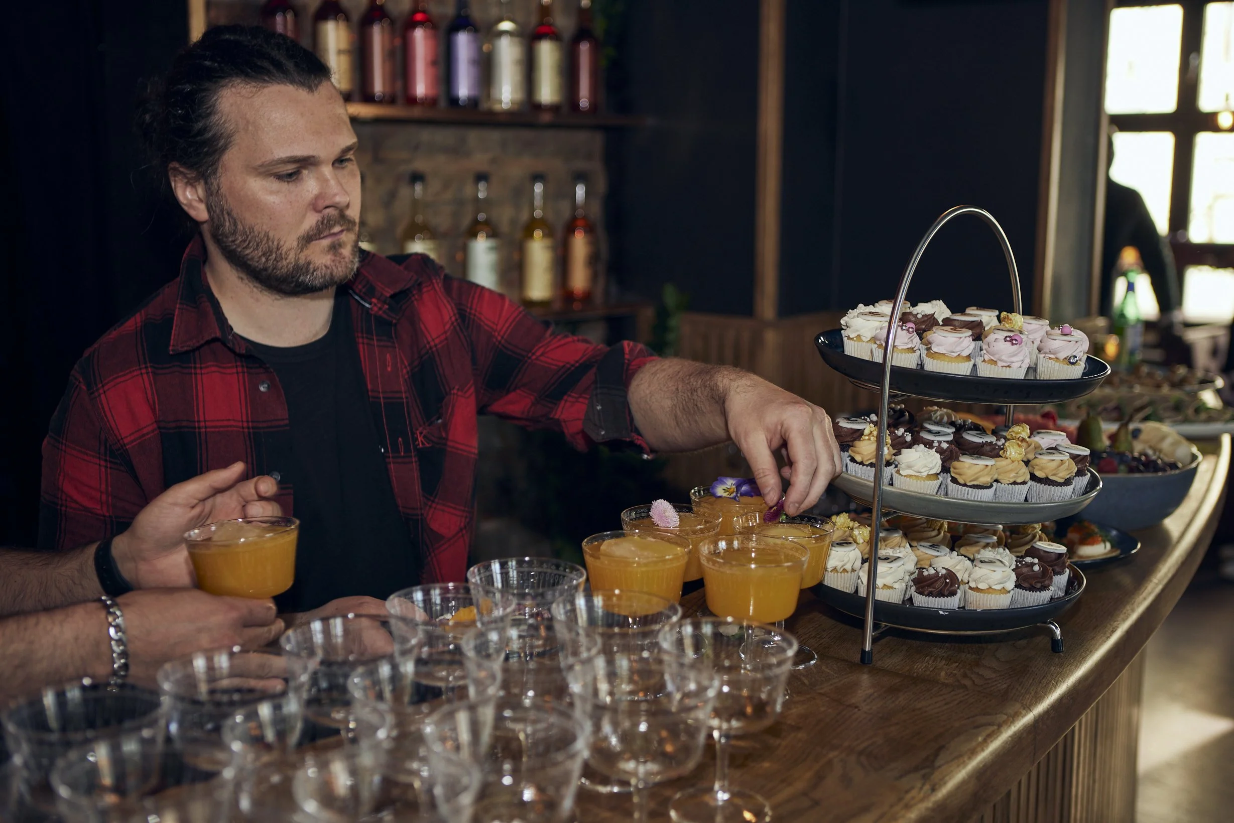 En mann med mørkt hår og skjegg, iført en rød og svart skinnskjorte, står foran en dessertbuffé med mange små cupcakes og glass med orange drikke. Man kan se flere personer rundt bordet, og bakgrunnen viser et kjøkken med hyller og sirupflasker.