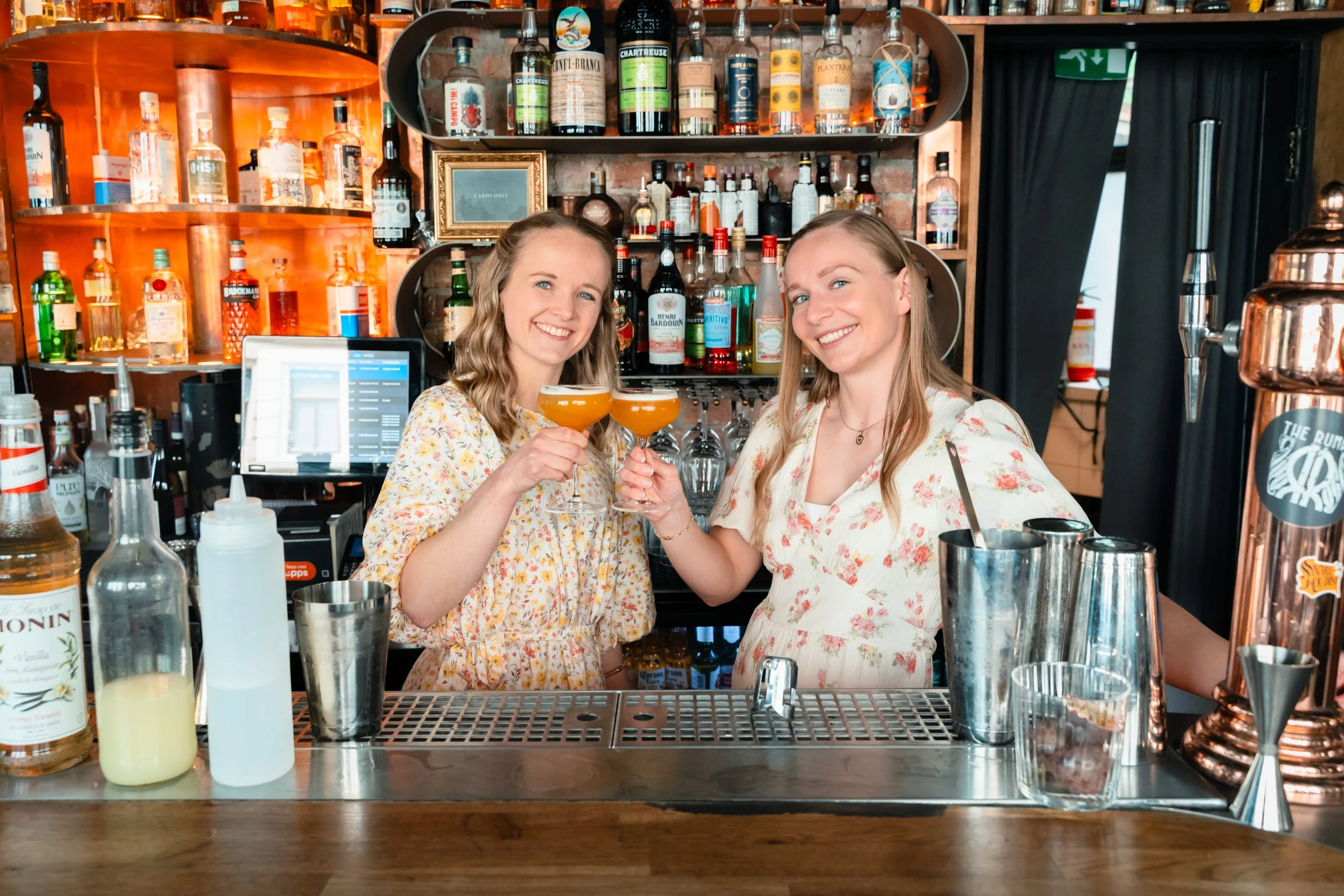 To kvinner smiler og har et glass med cocktail i en bar. Bakgrunn av hyller med ulike typer alkoholholdige drikkevarer.