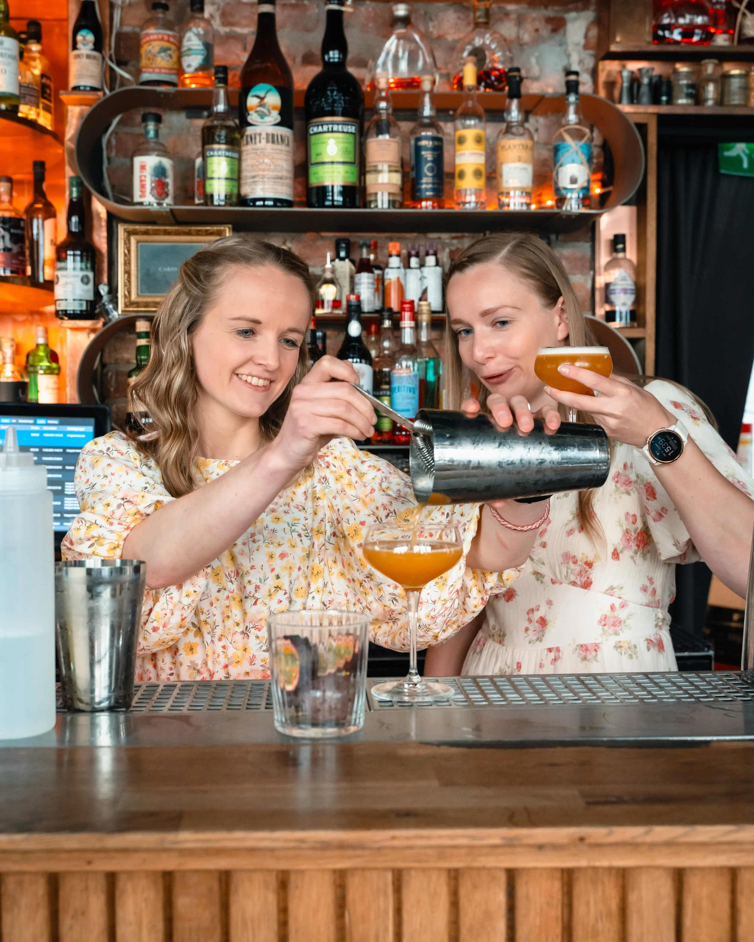 To kvinner som lager en drink på bartenderdisken med en ølglass i bakgrunnen