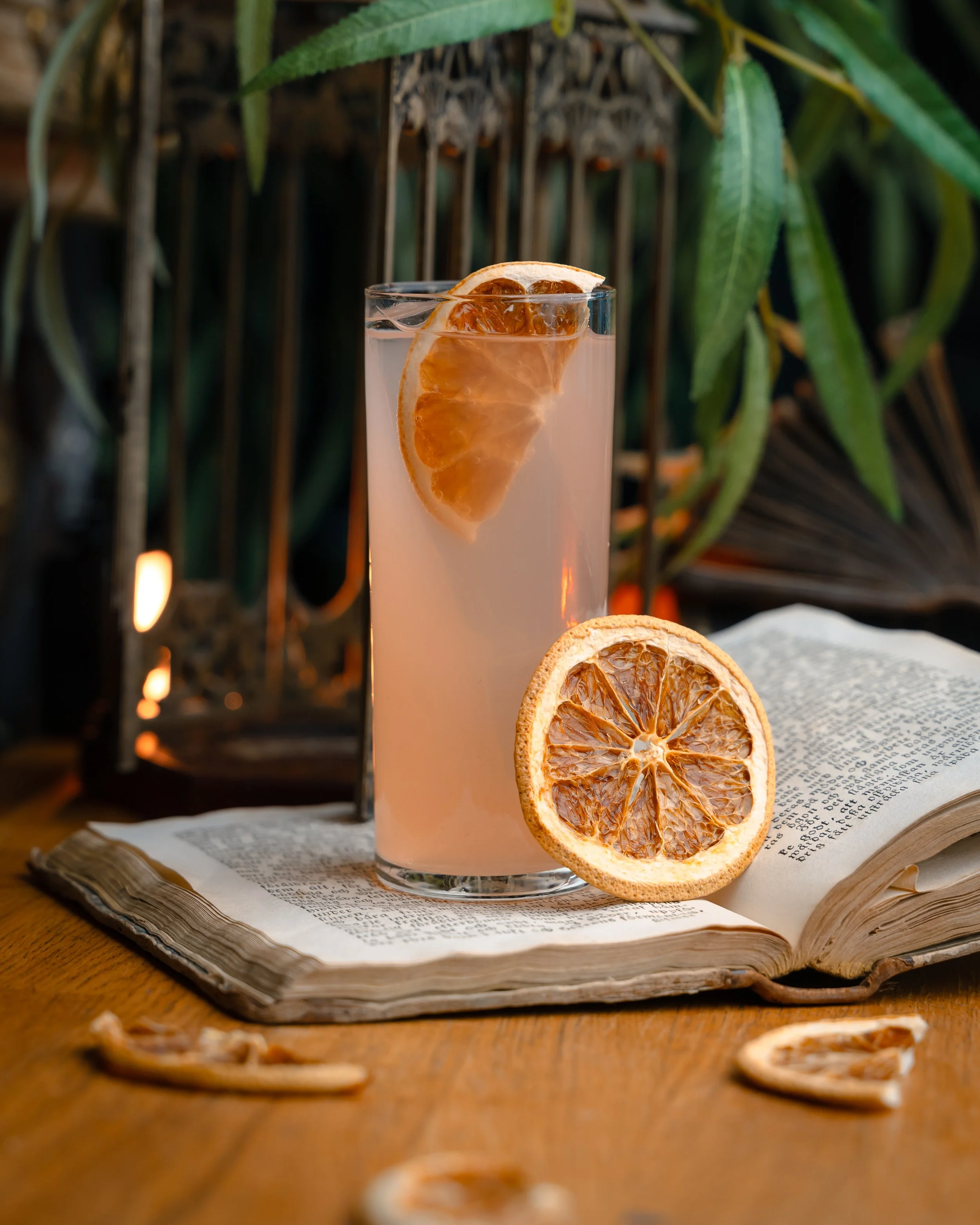 Et høyt glass med en sitrusdrink garnert med en skive sitron, plassert på en åpen bok med tekst, omgitt av tørkede sitrusbiter på en trebord.