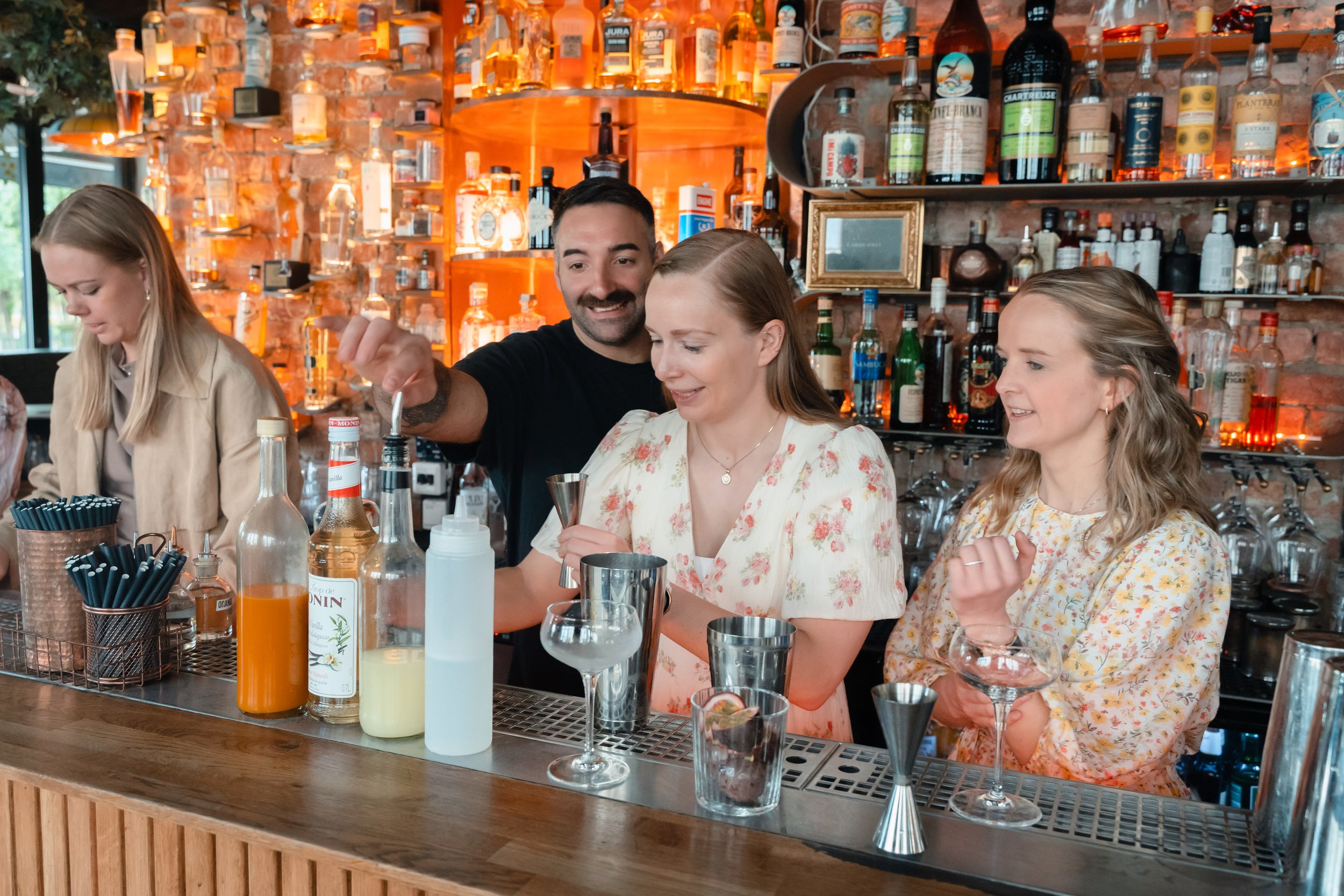 Fire personer som står bak en bar på en restaurant. En kvinne med lyst hår som holder en shaker, en mann med mørkt hår som smiler, og to kvinner med lyst hår som smiler. Baren har en murvegg med mange flasker med alkohol og glass. Det er ulike spritf