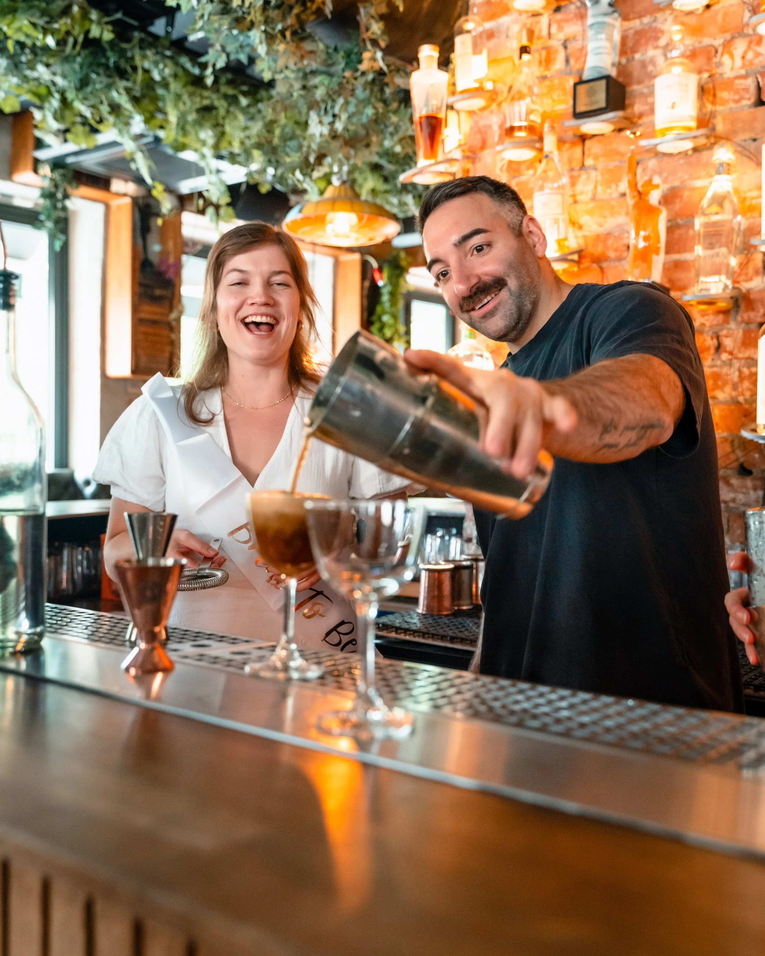 To personer bak en bar, en mann som heller en drink i et glass og en kvinne som ler, omgitt av glass, flasker og barutstyr, i en restaurant med brent murvegg og grønne planter i taket.