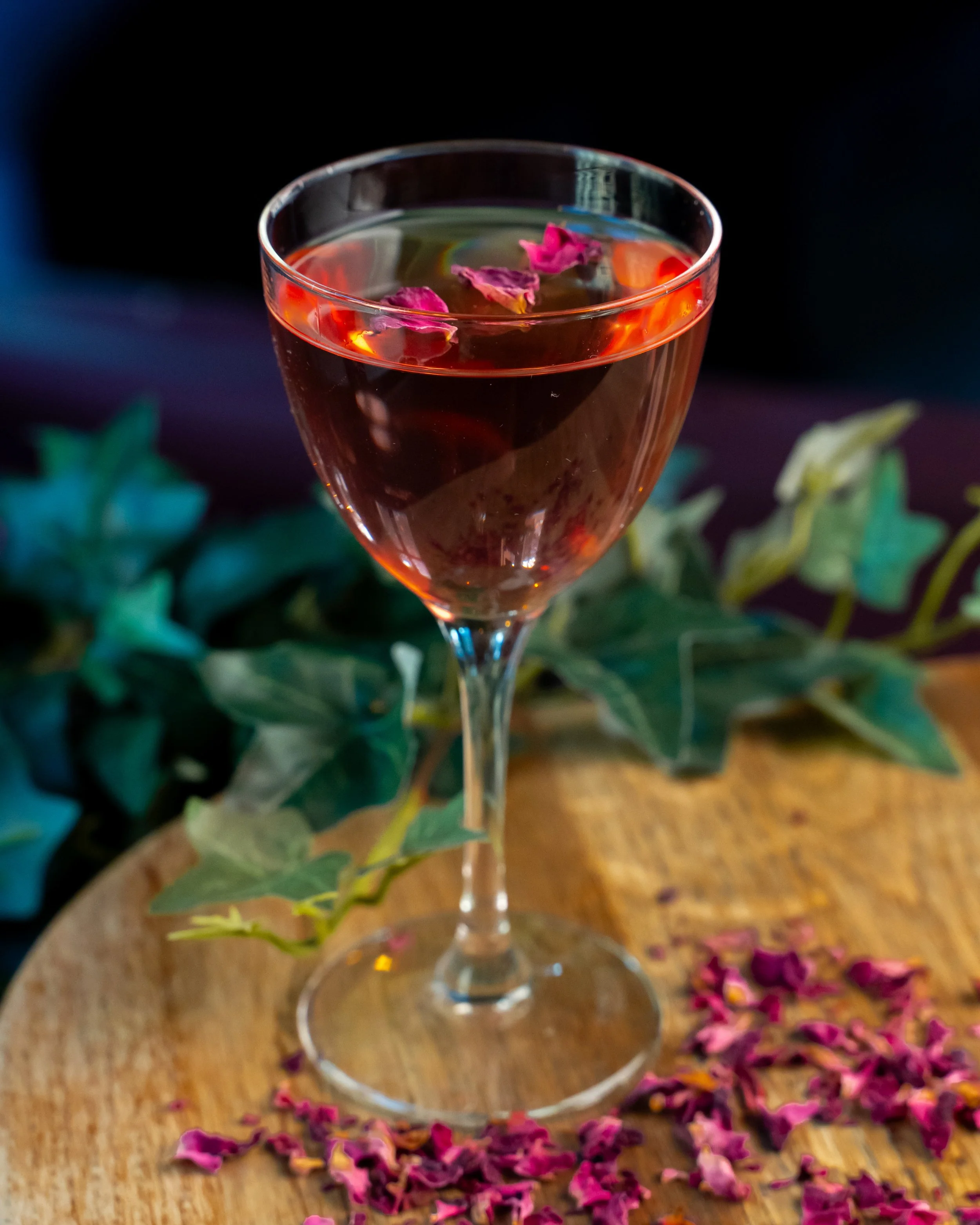 Et glass med rosa rosenblad og væske, plassert på en træbordplade, omgitt av flere rosa rosenblad på bordet.