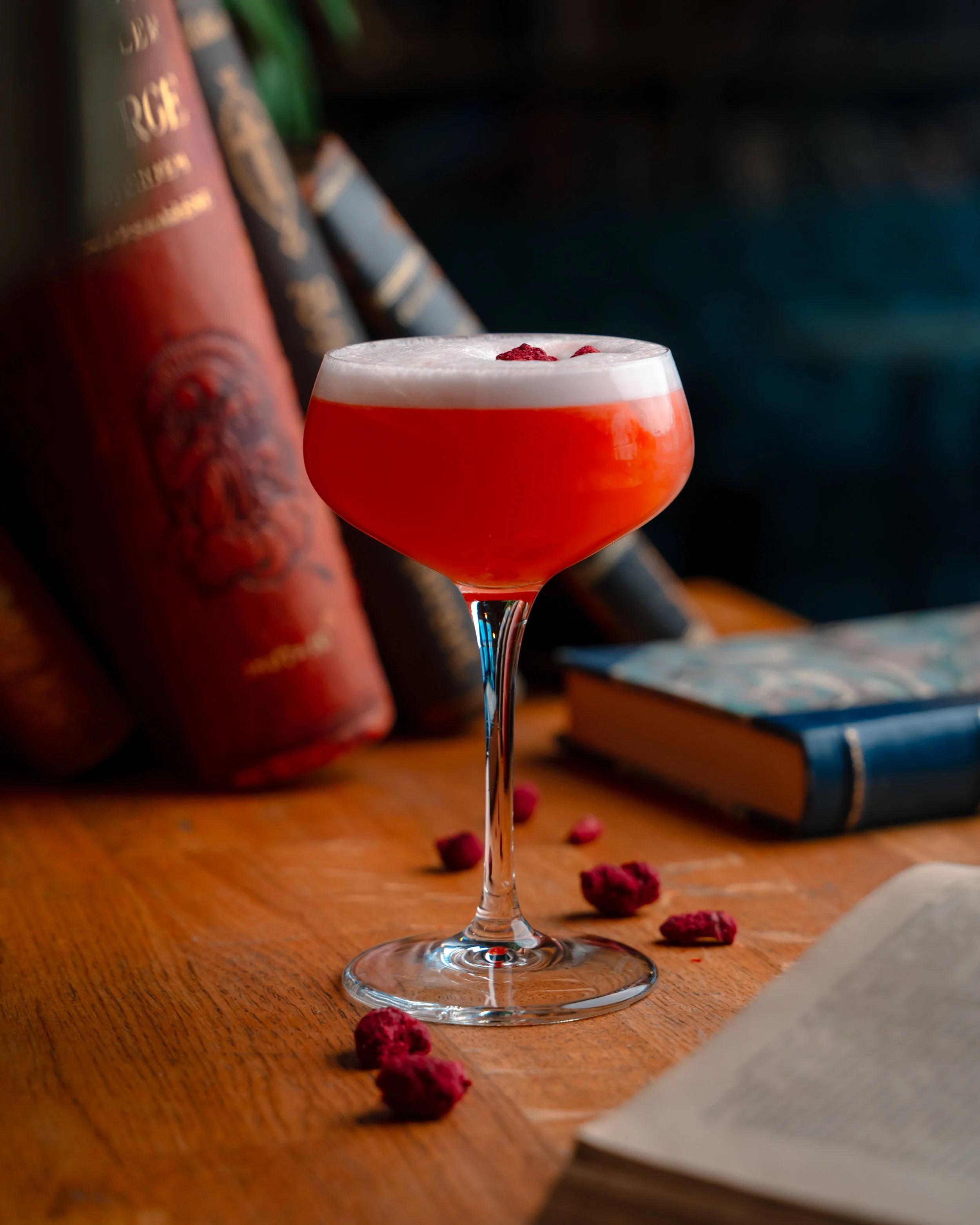 Et cocktail i et glass med en hvit skumtopper og små røde dekorasjoner, satt på en trebord med noen tørkede rosenblader spredt rundt.