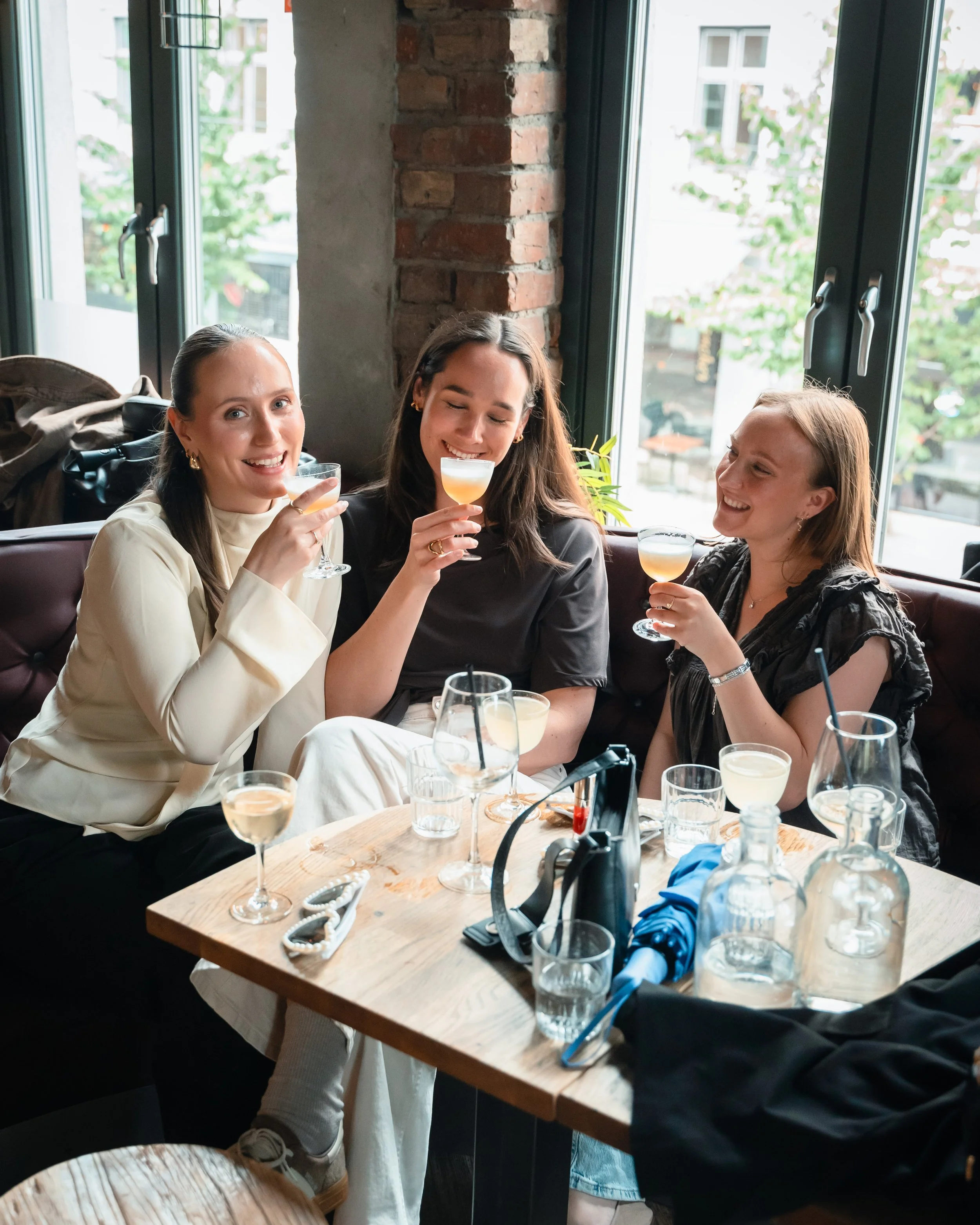 Tre unge kvinner som sitter rundt et bord på en restaurant eller kafé, og nyter drinker. De smiler og ser ut til å ha det hyggelig. Bordet er dekket med flere glass og noen gjenstander. Rommet har store vinduer og eksponert murvegg.
