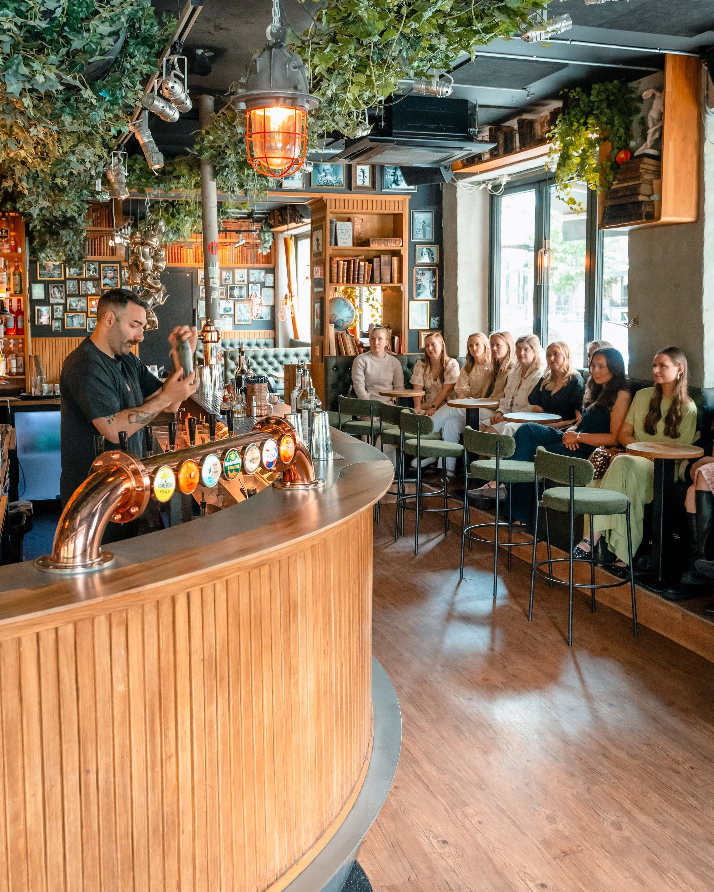 Interiør av en bar eller kafé med en bartender som gjør en drink, og en gruppe kvinner som sitter ved bord. Det er grønne planter og bilder på veggen, store vinduer som slipper inn naturlig lys, og atmosfæren virker koselig og innbydende.