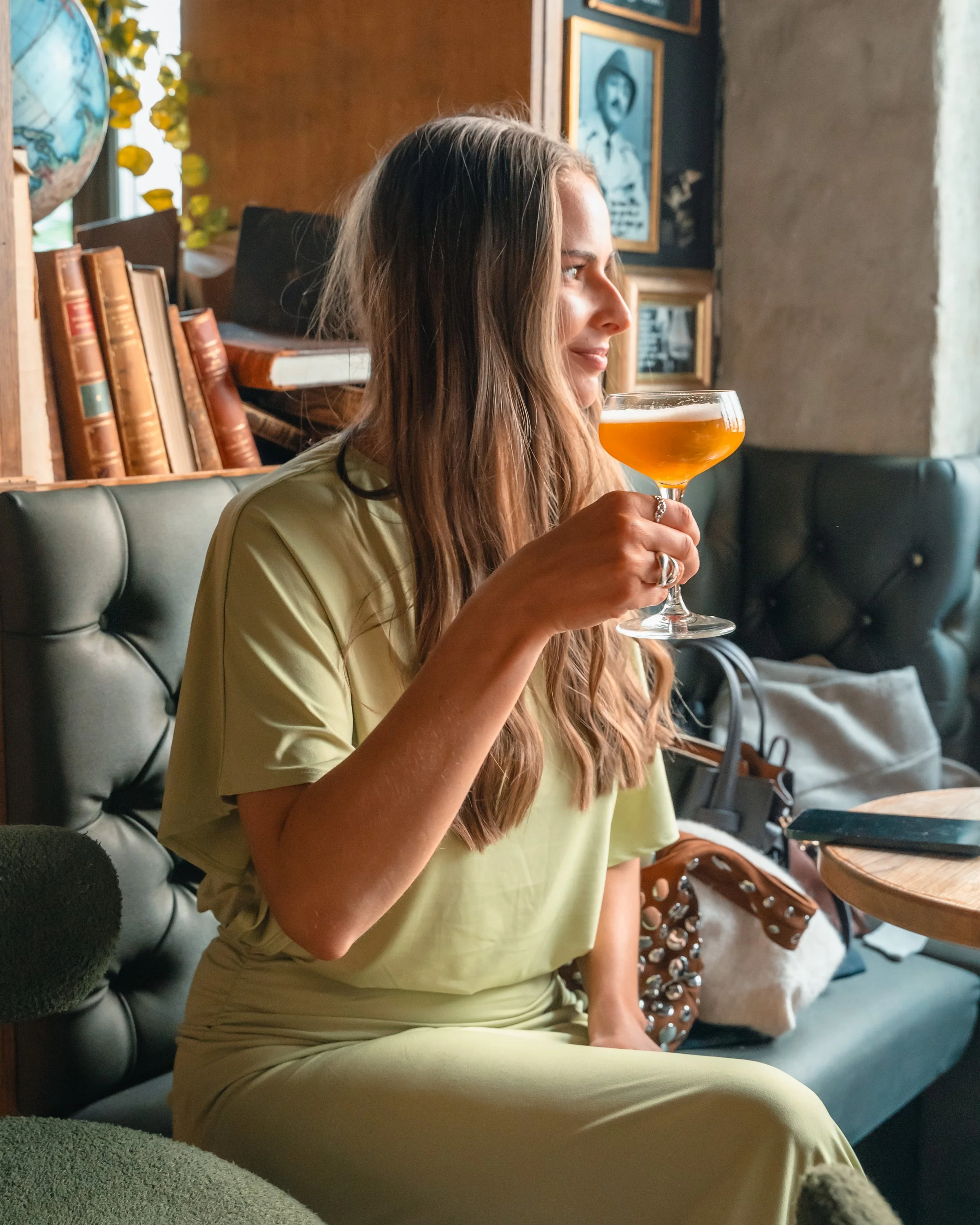 En ung kvinne med langt brunt hår smiler svakt mens hun holder et glass med øl. Hun sitter i en koselig kafe med en bokhylle og bilder på veggen i bakgrunnen, og det er grønne planter ved vinduet.