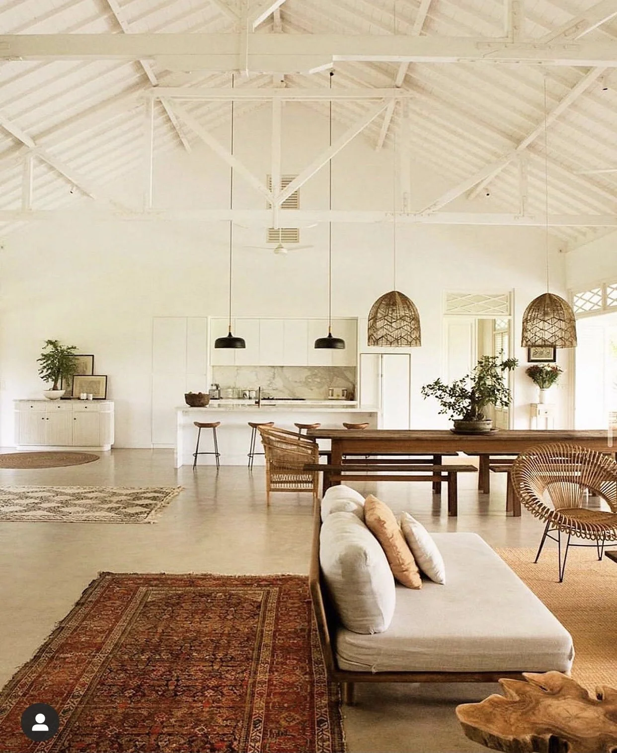 Un intérieur de maison lumineux avec un plafond en bois blanc, un canapé beige avec des coussins, des tapis et des meubles en bois, une cuisine en arrière-plan avec un îlot, deux suspensions en bambou et des plantes décoratives.