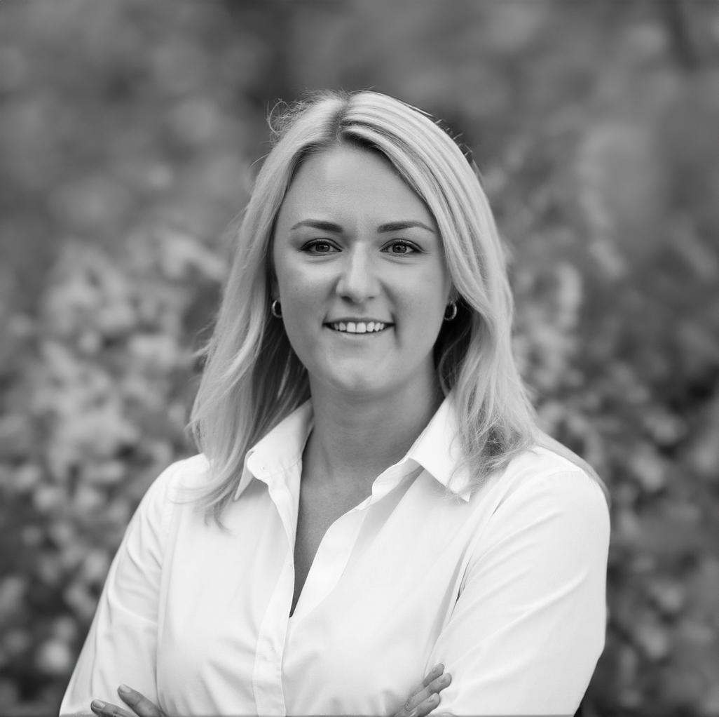 Portrait d'une femme blonde souriante, portant une chemise blanche, devant un fond flou de paysages naturels.