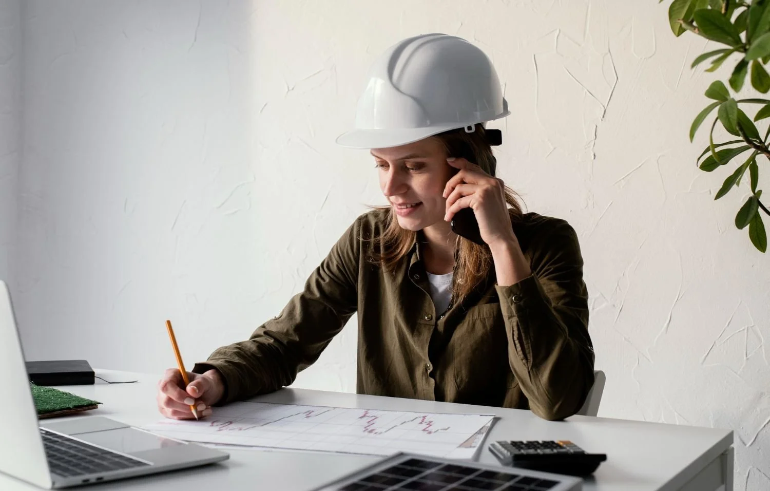 Woman wearing a safety helmet talking on the phone and working on architectural plans at a desk.