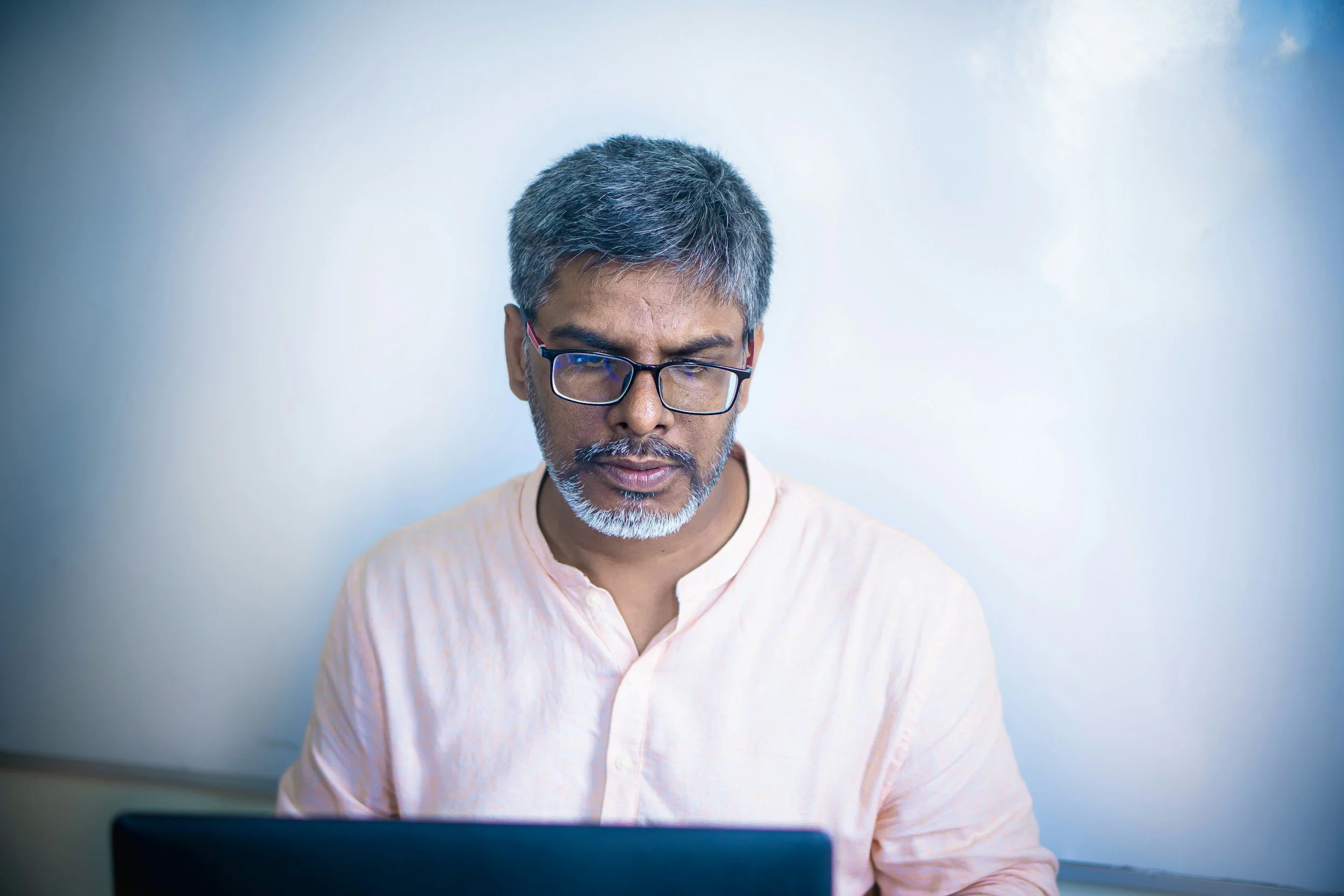 Mid-aged man with short gray hair and beard wearing glasses and a light pink shirt looking down at a laptop in front of him.