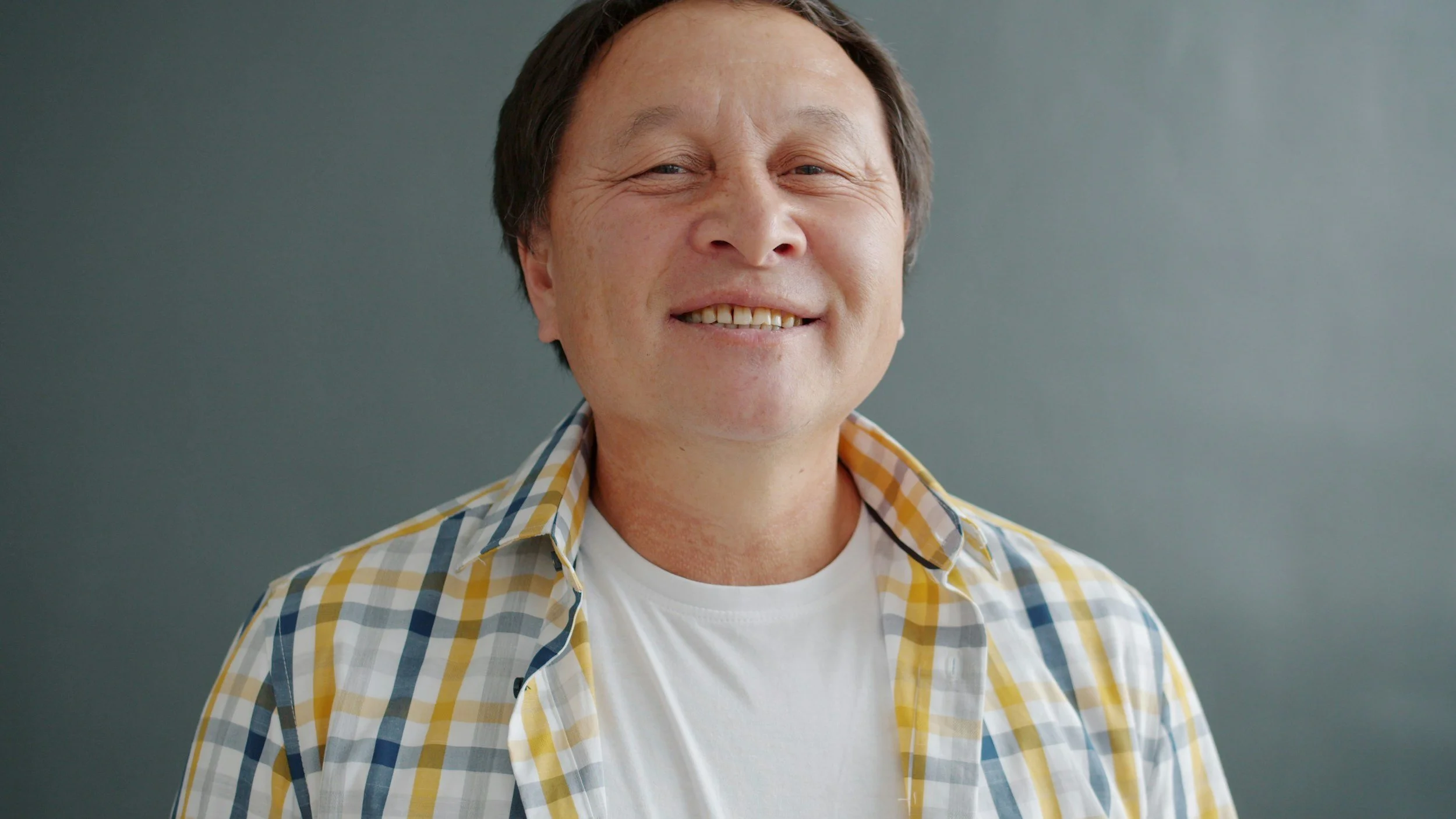Smiling man with short dark hair wearing a yellow and blue checked shirt over a white t-shirt, standing against a plain wall.