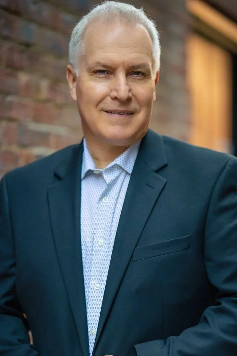 A middle-aged man with gray hair dressed in a dark suit and light patterned shirt, standing outdoors in front of a brick wall, smiling confidently at the camera.