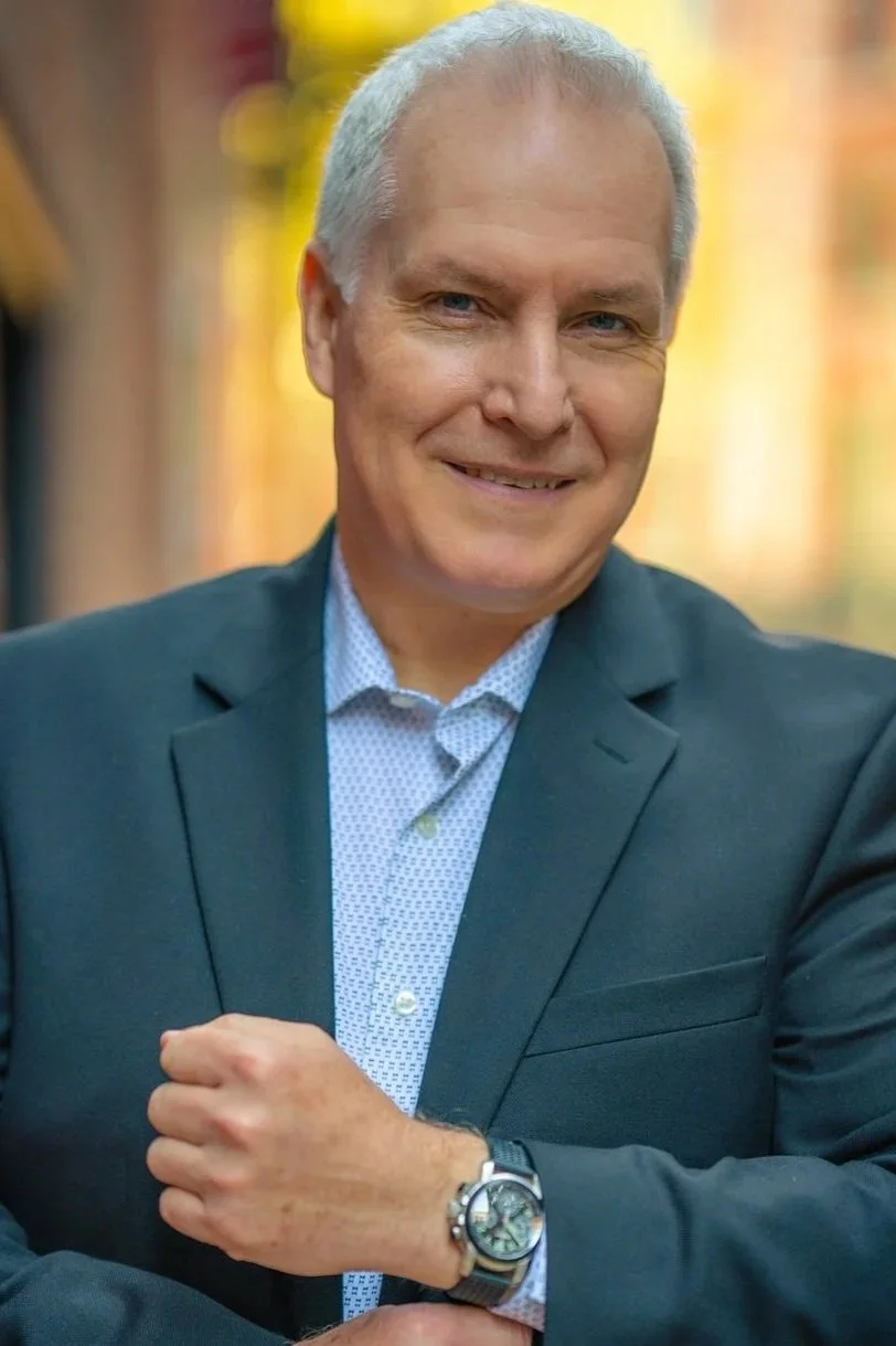 A middle-aged man with silver hair, dressed in a dark suit and light patterned shirt, smiling at the camera. He is wearing a watch on his left wrist and is standing outdoors with blurred warm-colored background.