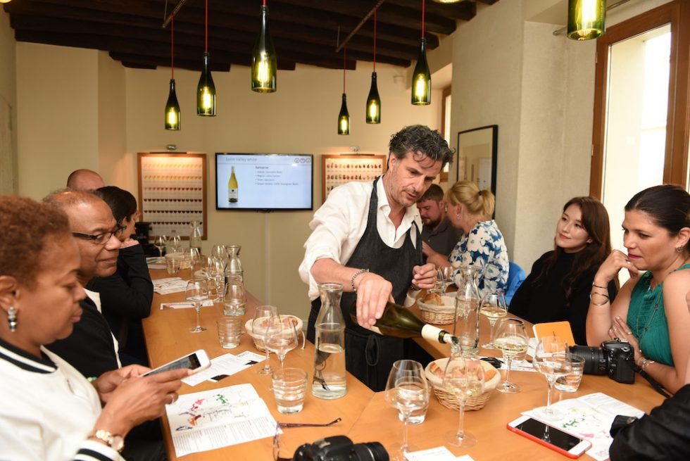 A chef demonstrates pouring wine at a wine tasting event in a room with a long table, diverse participants, and a digital screen in the background.