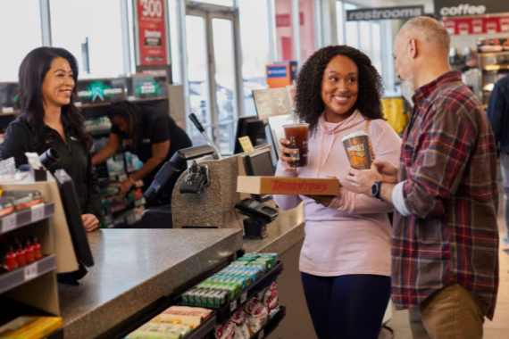 A woman and a man smiling and holding drinks at a convenience store checkout counter, talking, with a store employee in the background.